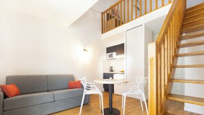 A modern living room with a grey sofa, white chairs, a round table, wooden staircase, and a kitchen area with white cabinets. - City Résidence Bordeaux Centre (Bordeaux)