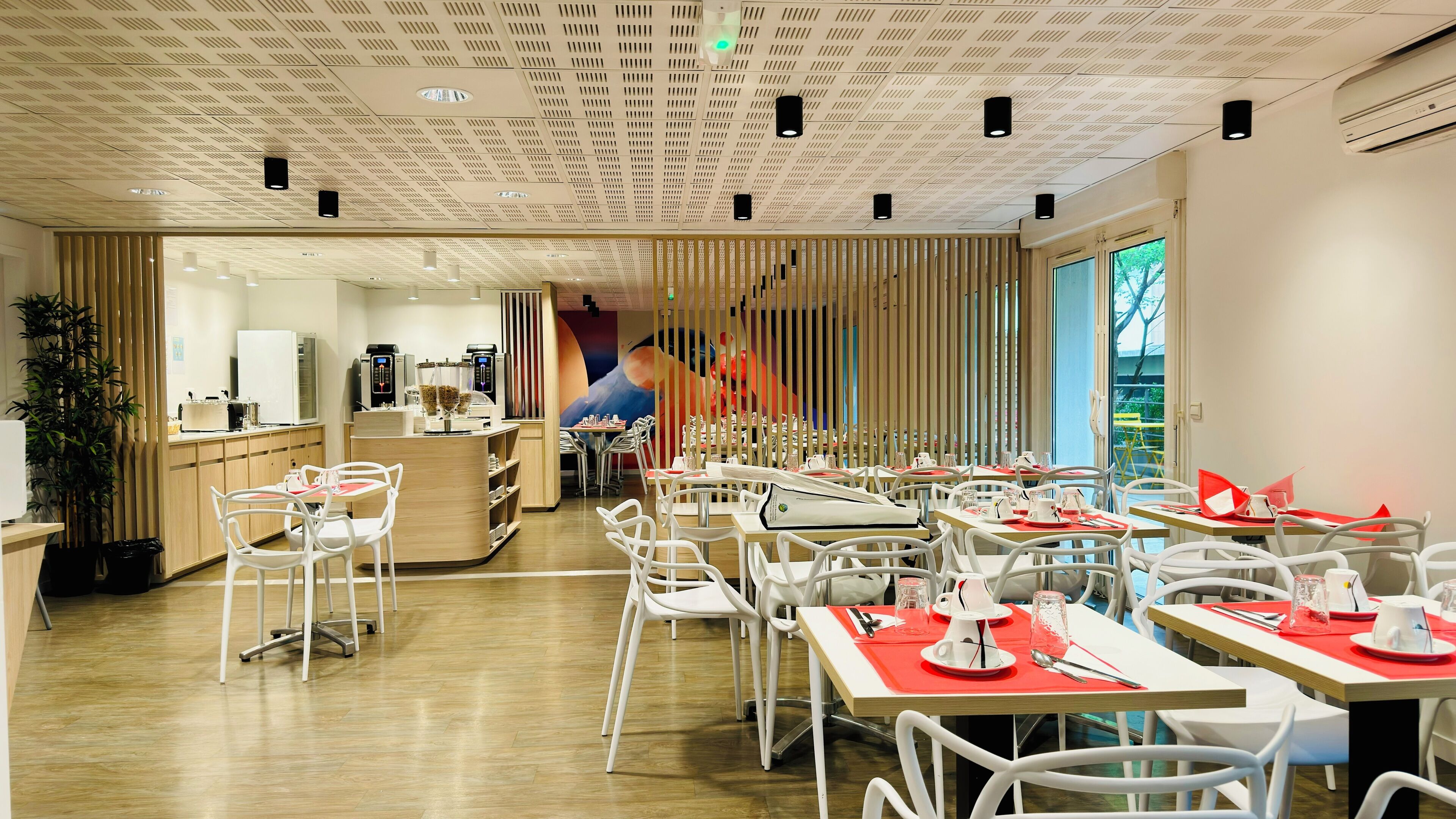 Een modern restaurant met witte stoelen en tafels, een centraal gelegen toonbank met koffiemachines, en een grote muurschildering op de achterwand.