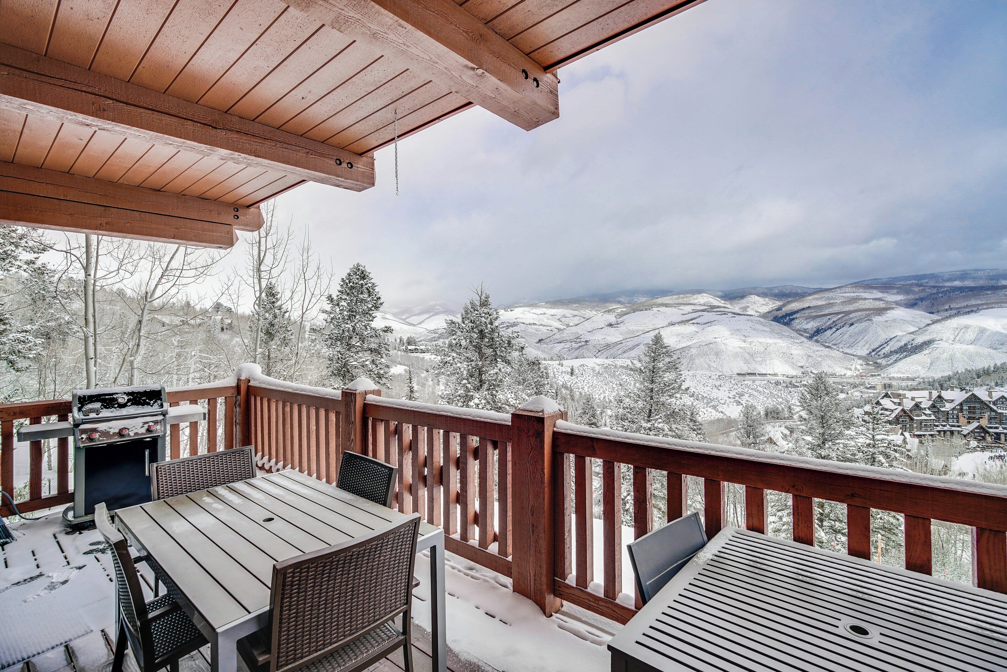 A wooden deck with a table and chairs, overlooking a snowy landscape with mountains.