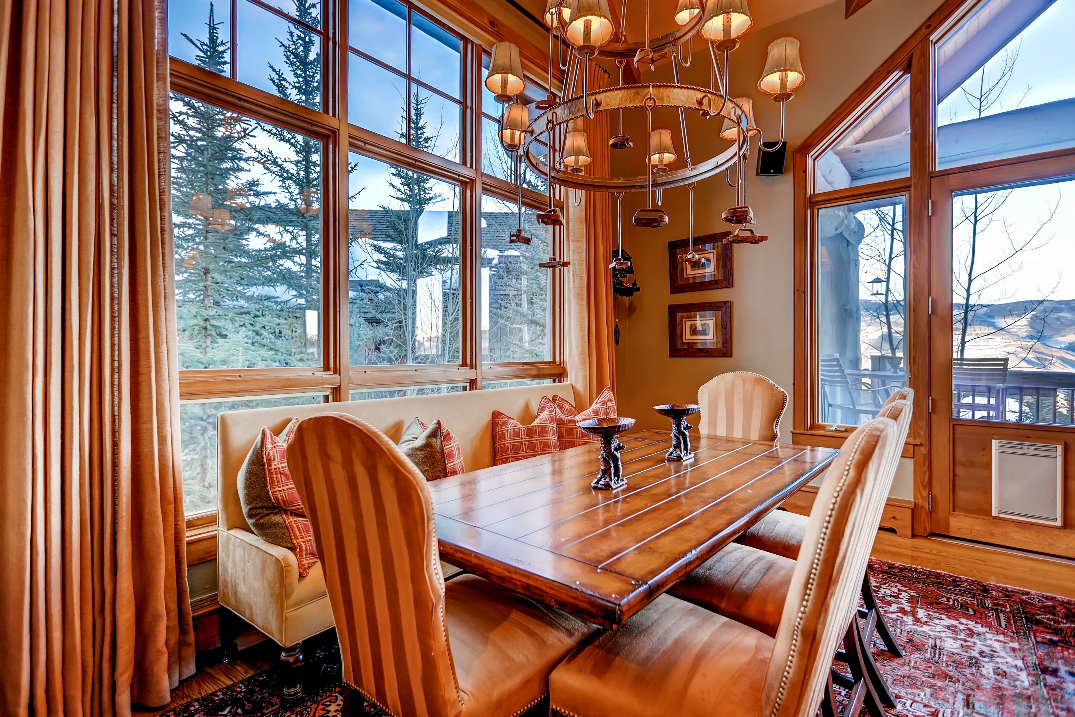 A dining area with a wooden table and leather chairs, large windows with a view of trees, and a chandelier.