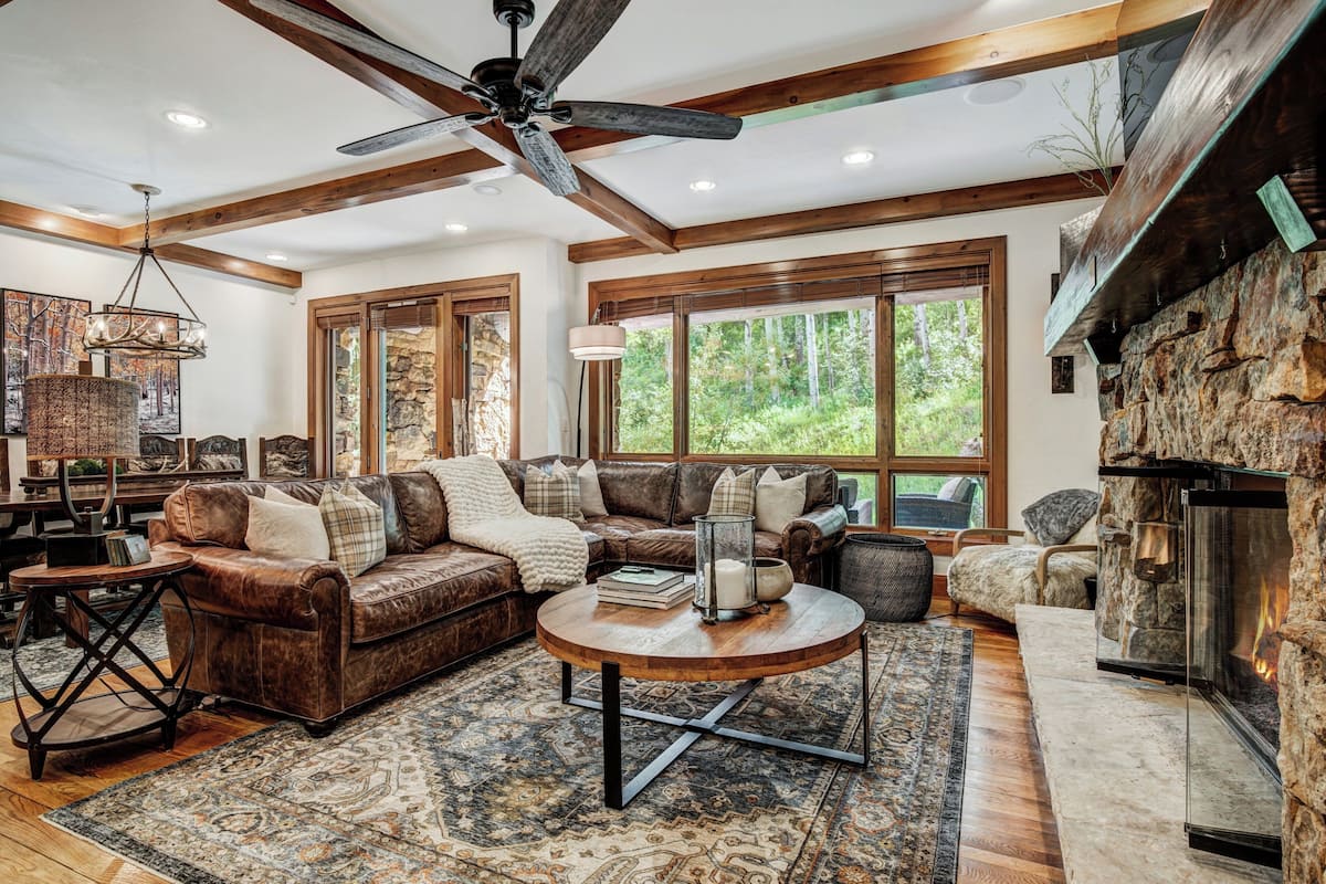 A spacious living room with a fireplace, large windows, and a ceiling fan.