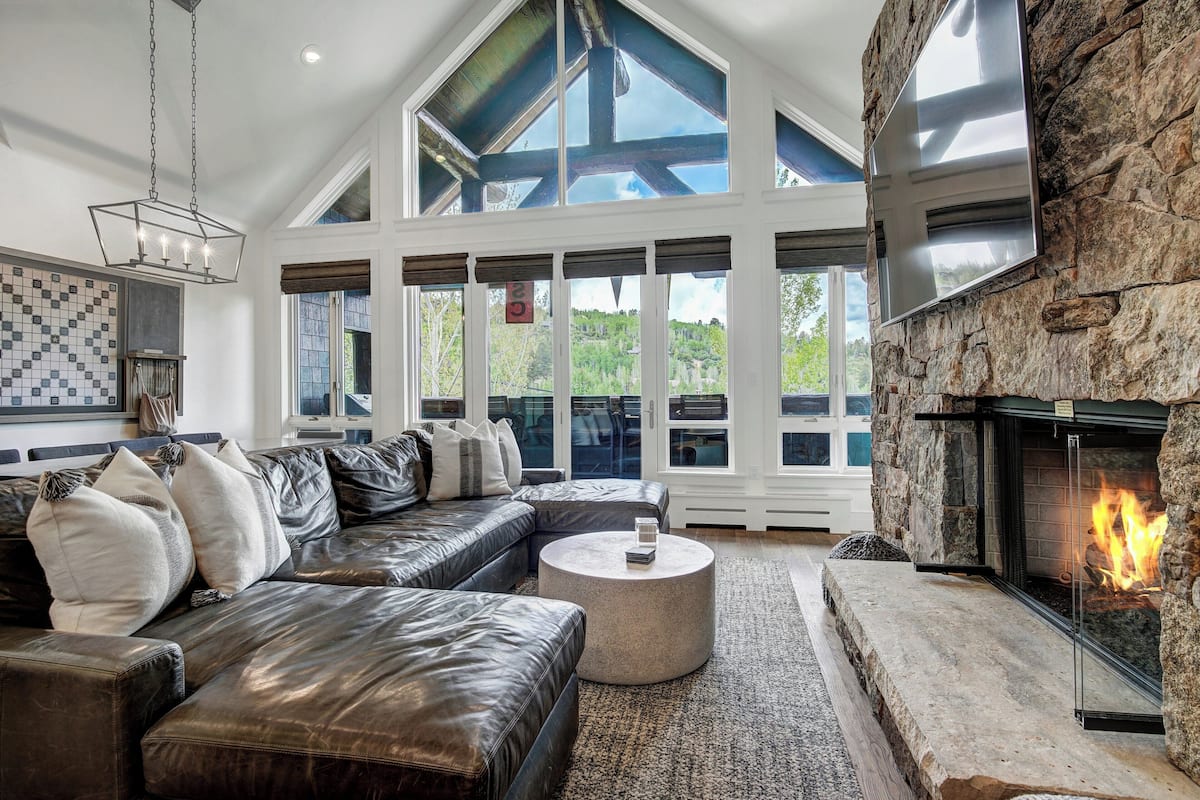 A spacious living room with a stone fireplace, leather sofas, and large windows offering a view of the outdoors.