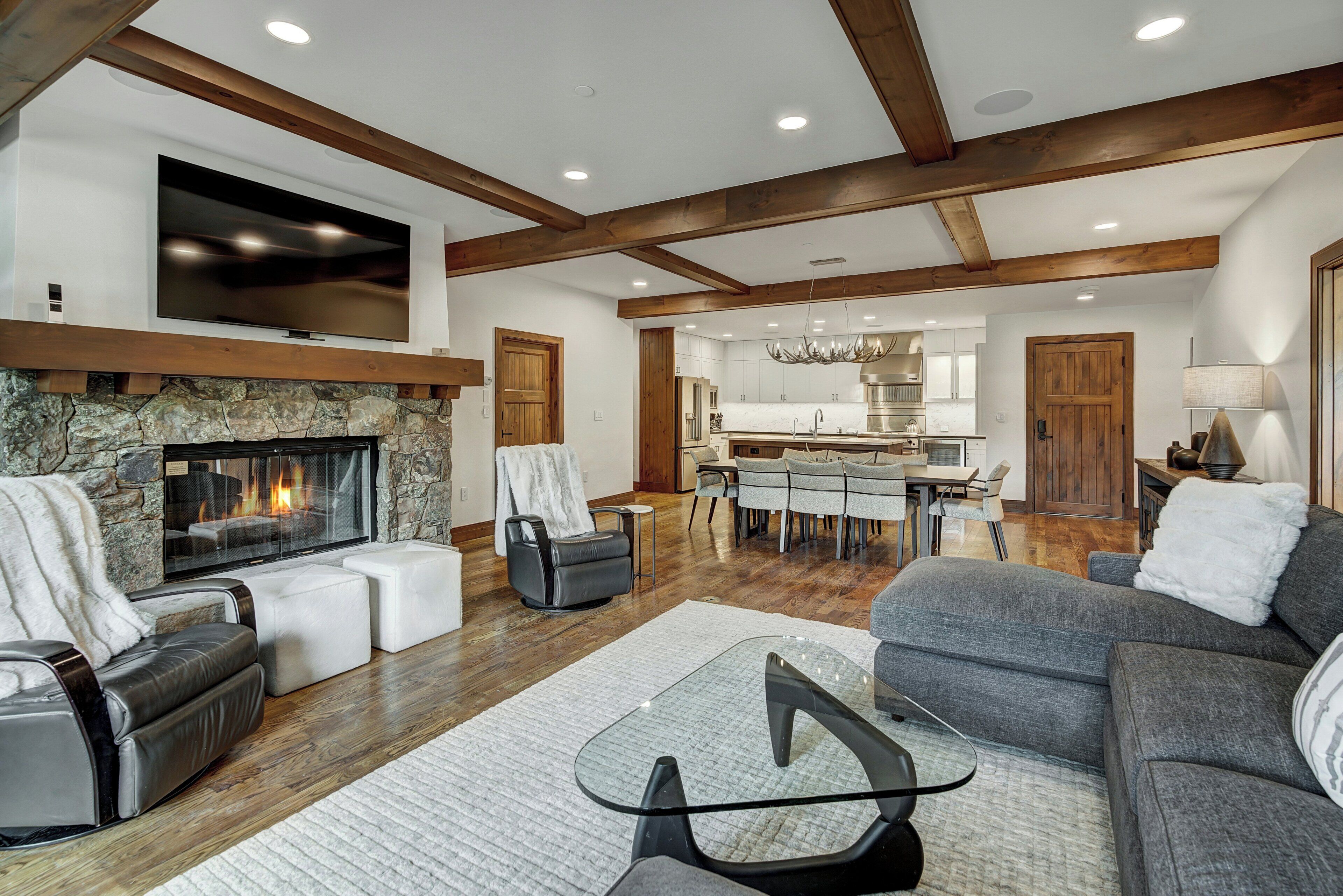 A spacious living area with a stone fireplace, a flat-screen TV mounted above it, and a modern kitchen in the background.