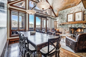 A spacious dining area with a large wooden table and chairs, large windows offering a view of the outdoors, and a stone fireplace with framed artwork above. - Bachelor Gulch Village by Vail Resorts (Avon)