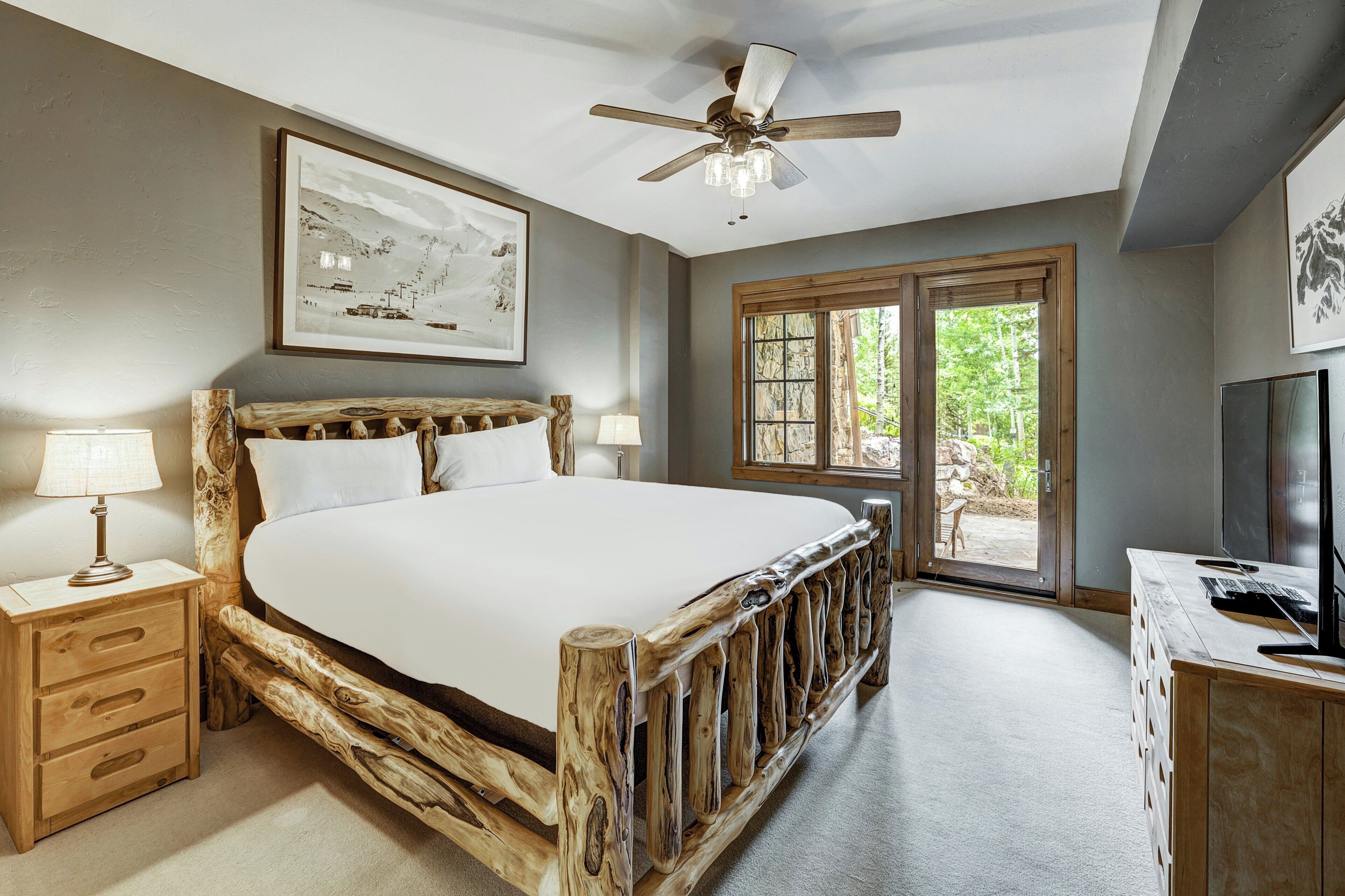 A bedroom with a rustic wooden bed frame, a ceiling fan, a large window with a view of greenery, and a television on a wooden dresser.