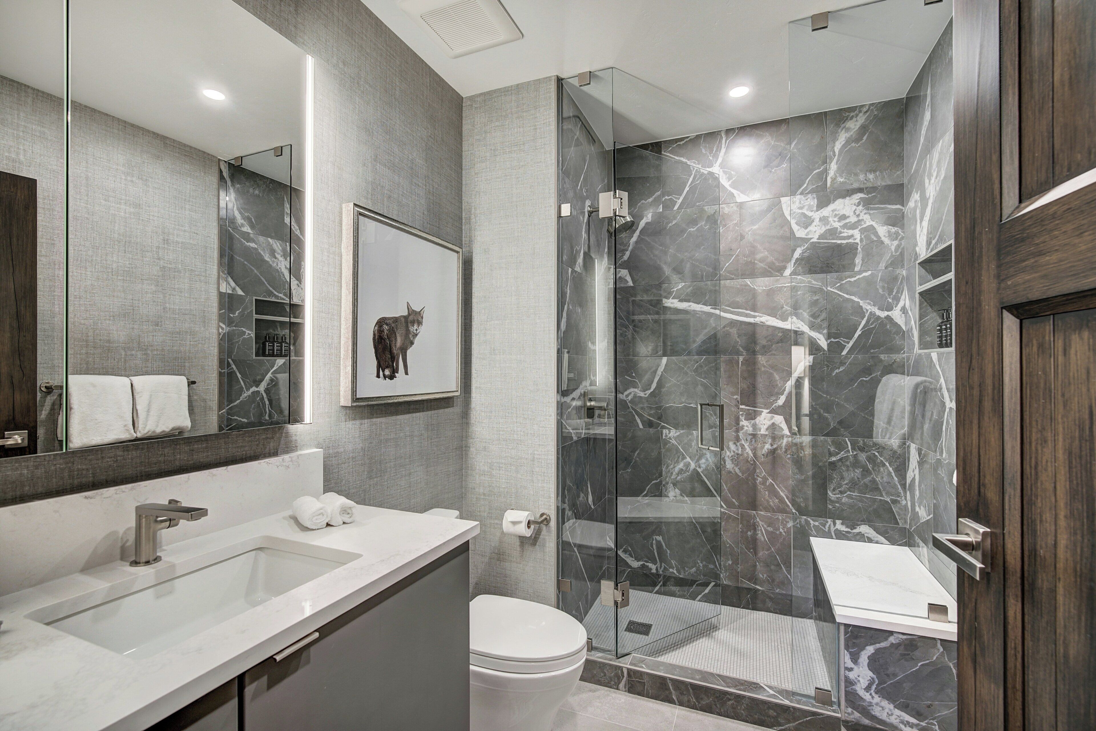 A modern bathroom with a large mirror, a marble shower wall, and a white sink.