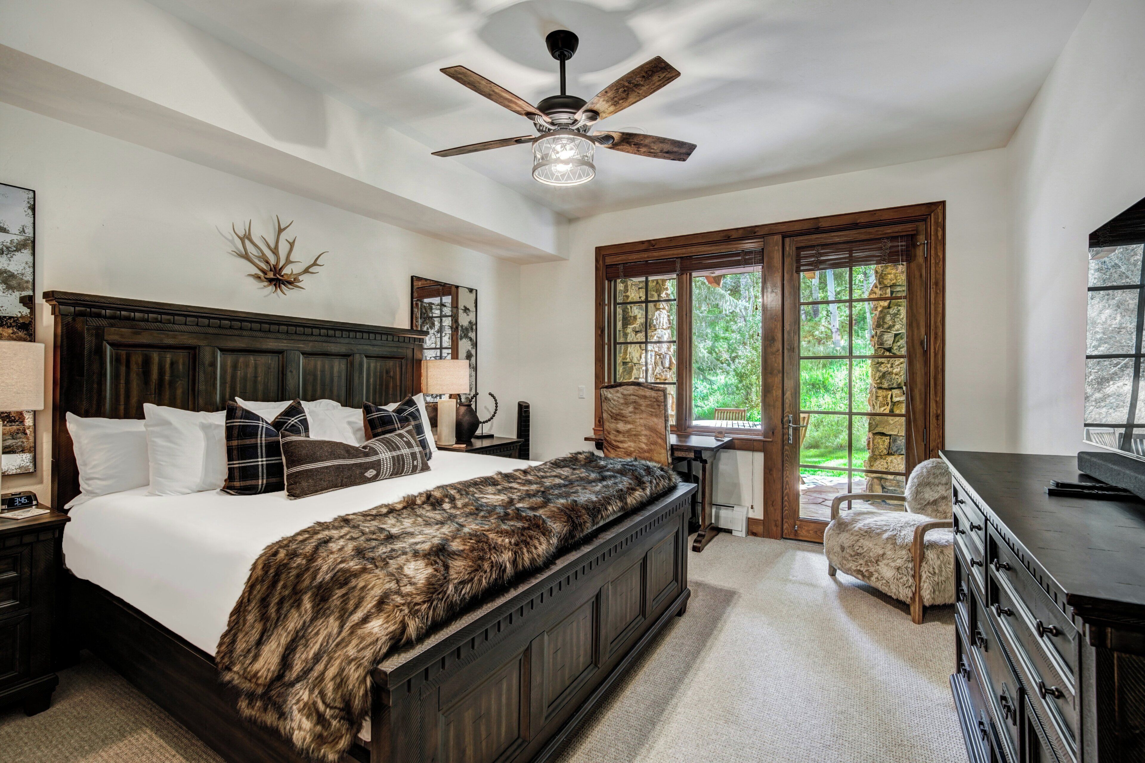 A bedroom with a large bed, a ceiling fan, a stone fireplace, and a view of the outdoors through a glass door.