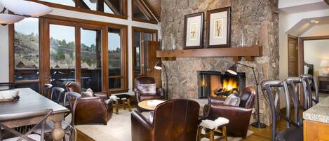 A spacious living room with a stone fireplace, wooden beams, and large windows offering a view of the outdoors.