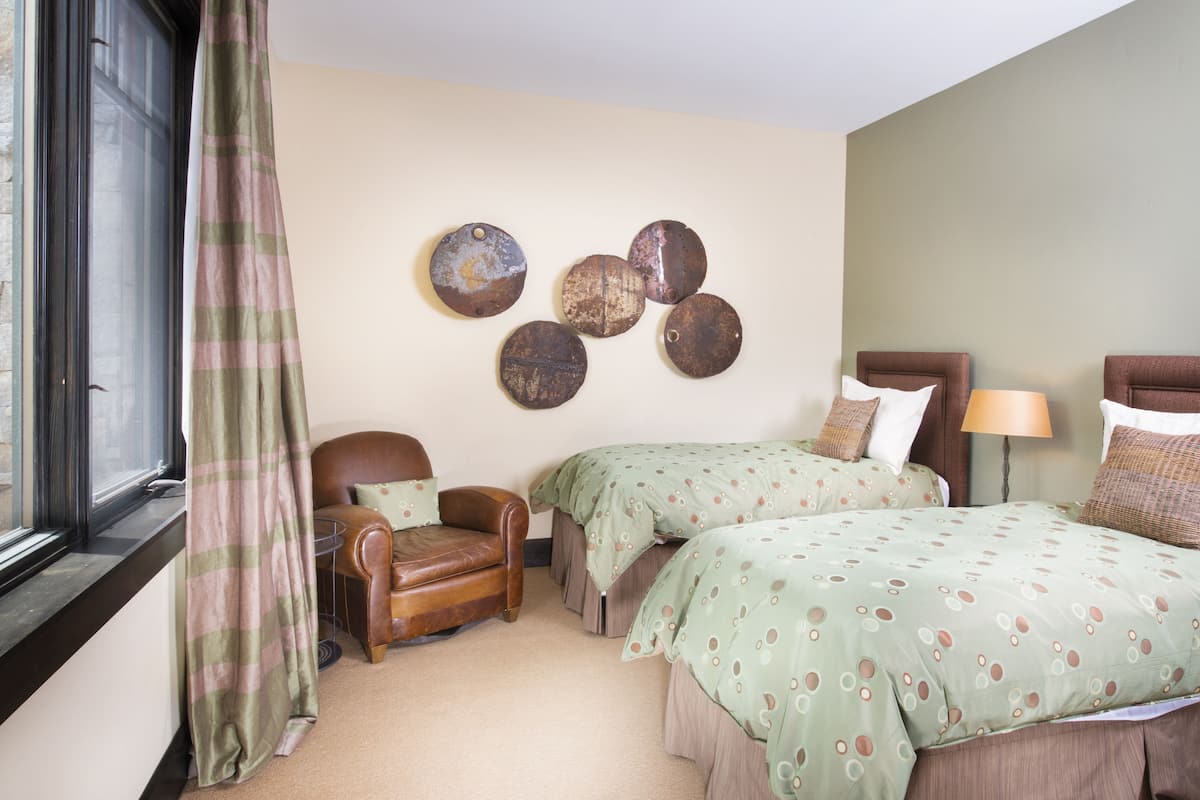 A hotel room with two beds, a chair, a lamp, and a window with a curtain.