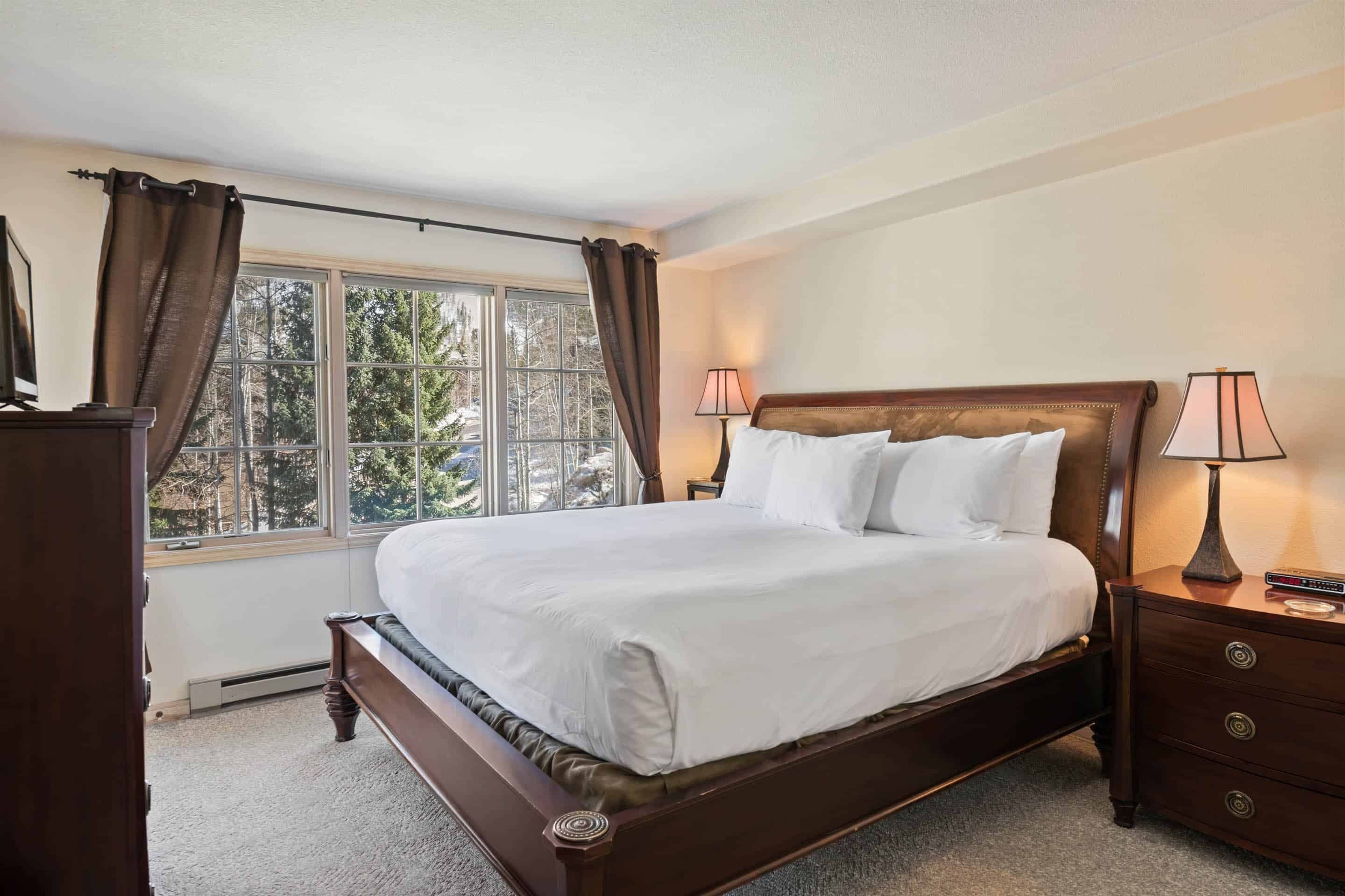 A neatly made bed in a hotel room with a large window, two bedside lamps, and a wooden nightstand.