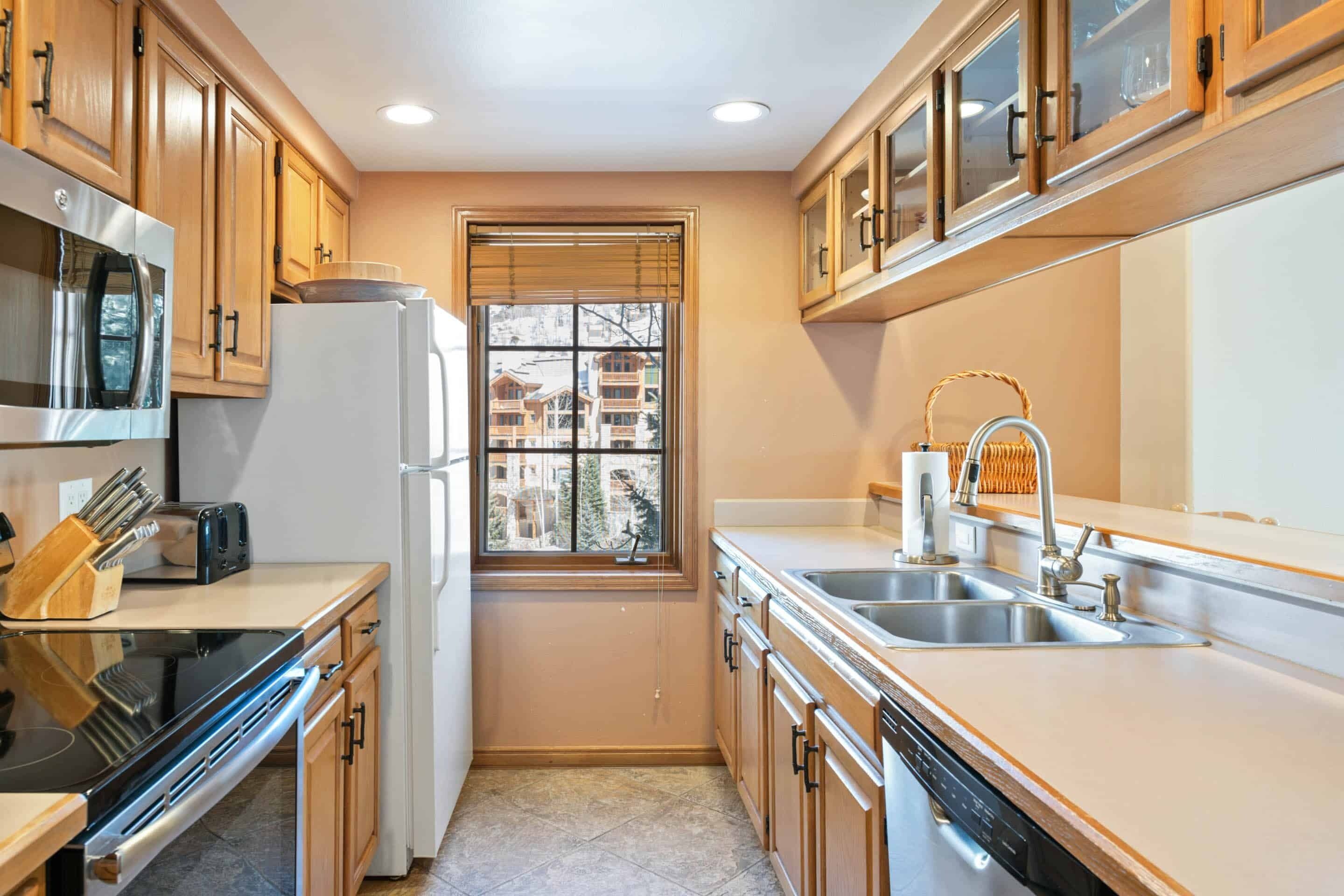 A compact kitchen with wooden cabinets, a built-in microwave, a refrigerator, a sink, and a window with blinds.