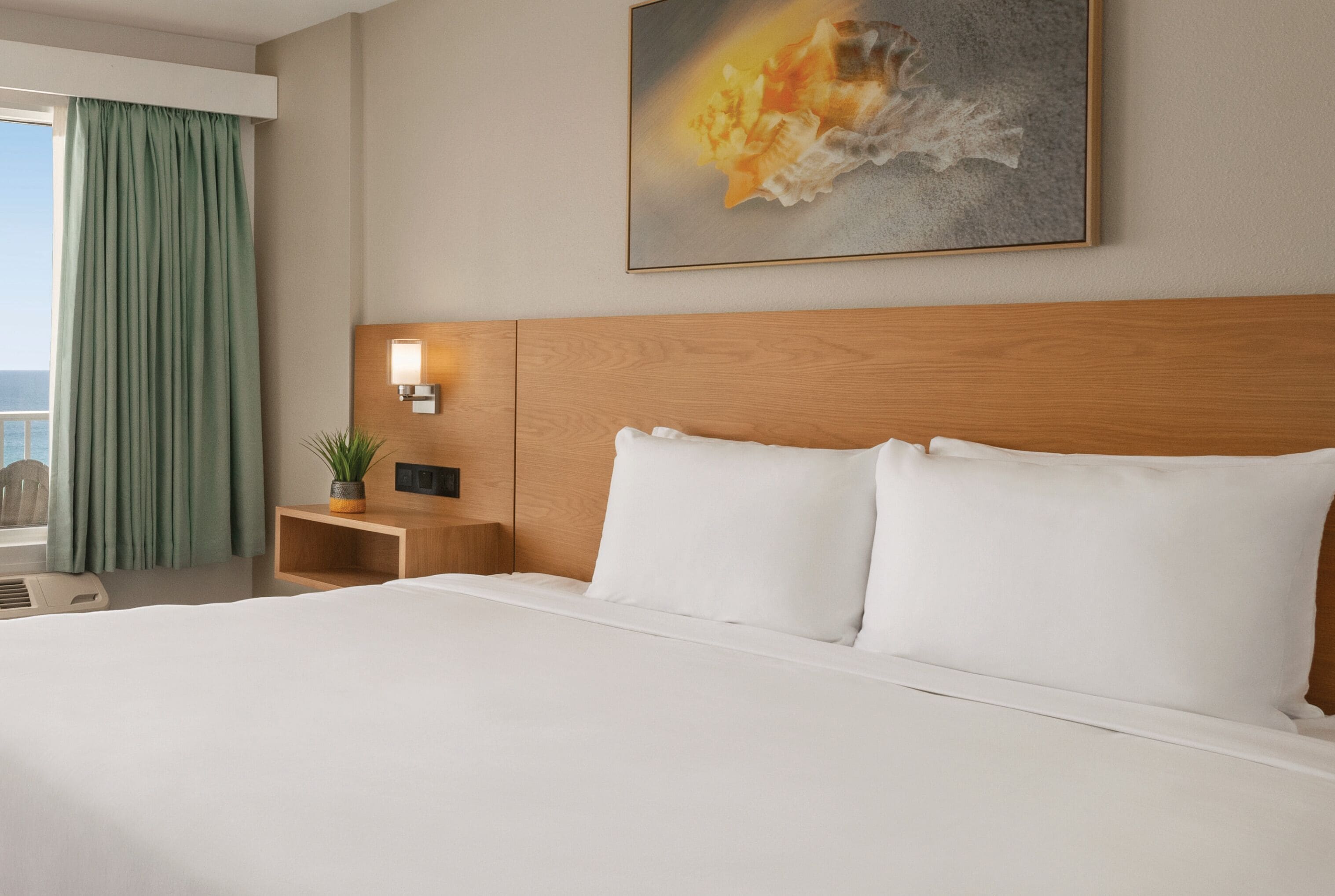 A hotel room with a large bed, a wooden headboard, a nightstand with a lamp, a potted plant, and a painting on the wall.