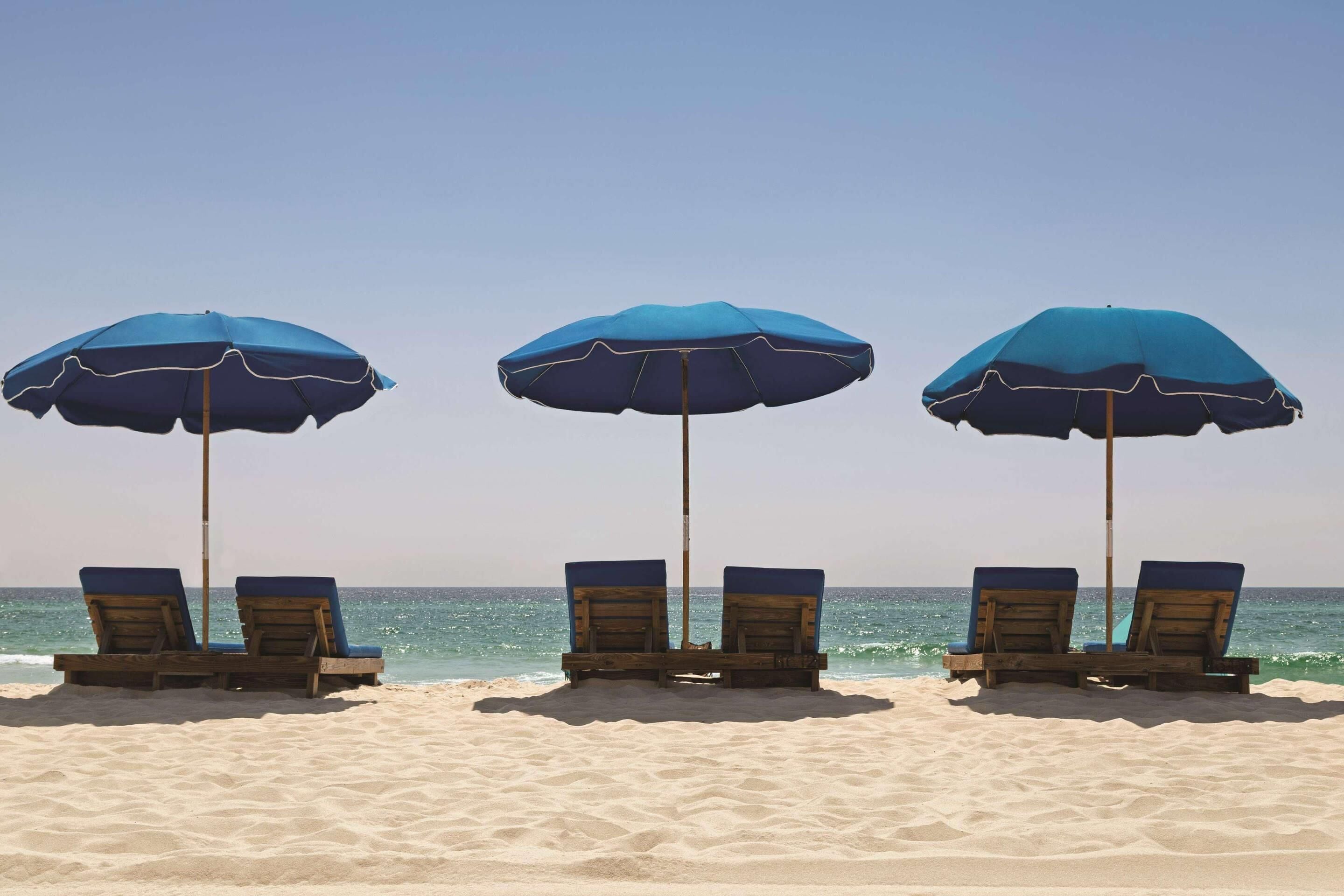 Drei Strandstühle und Sonnenschirme auf einem Sandstrand, im Hintergrund das Meer.