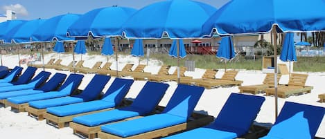 Filas de sillas de madera en la playa con cojines y sombrillas azules.