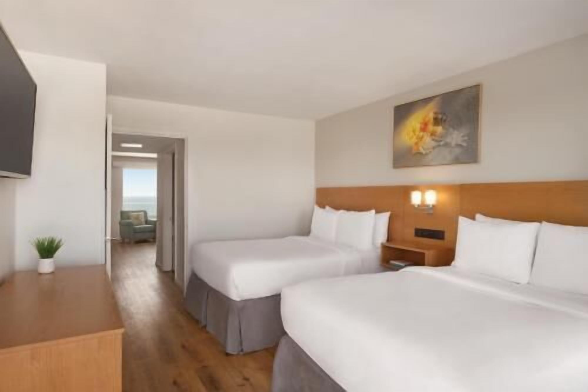 A hotel room with two beds, a TV, a painting, and a view of the sea.