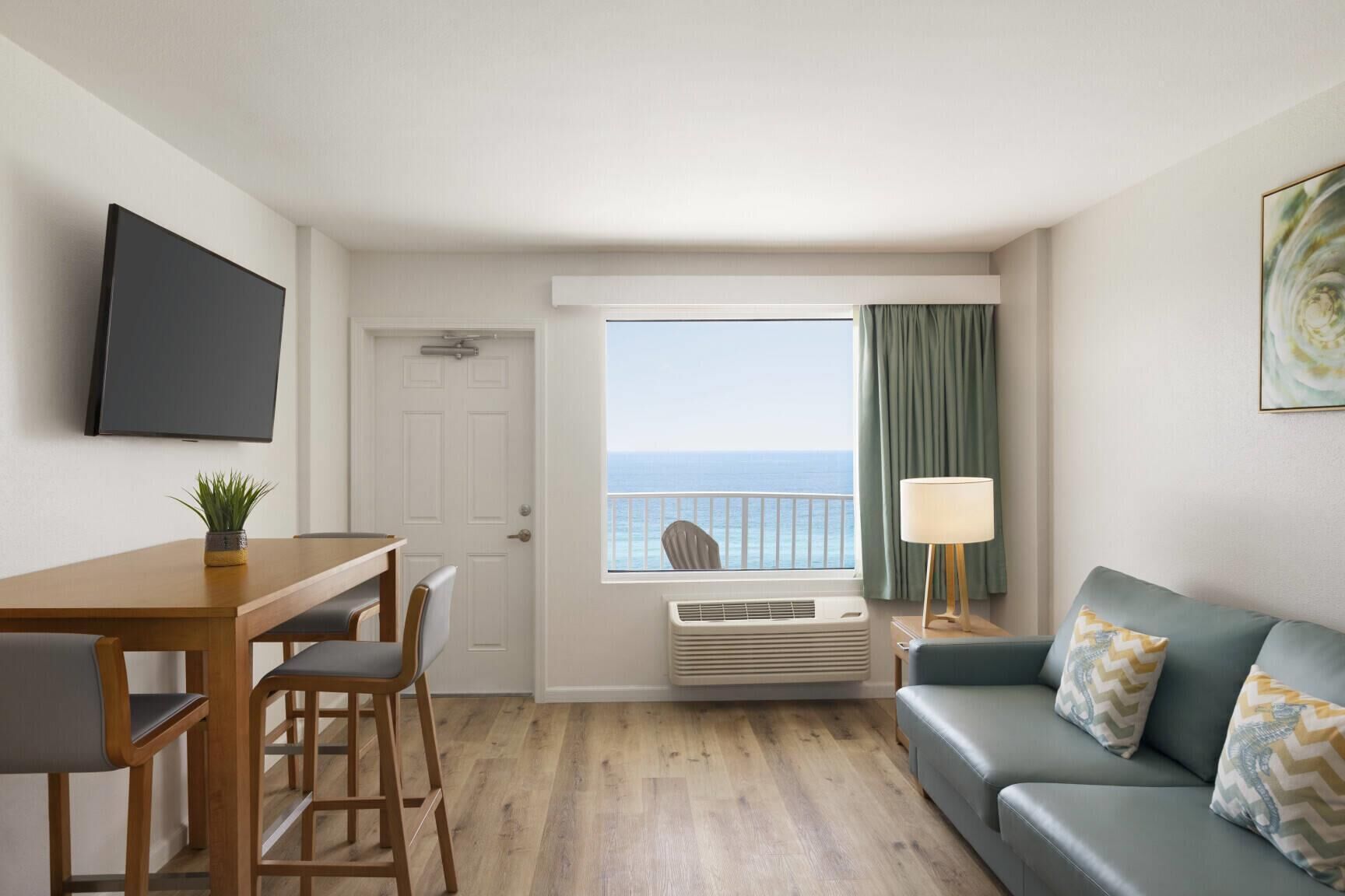 Una habitación con una mesa de madera, dos sillas, un sofá, un televisor montado en la pared, un ventanal con vista al mar y un ventilador de pie.