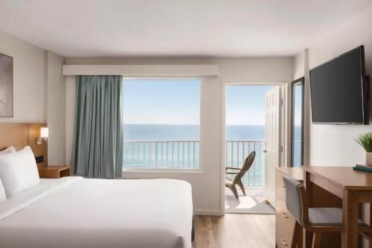 Een hotelkamer met een bed, een bureau, een stoel, een televisie, een raam met uitzicht op zee en een balkon.