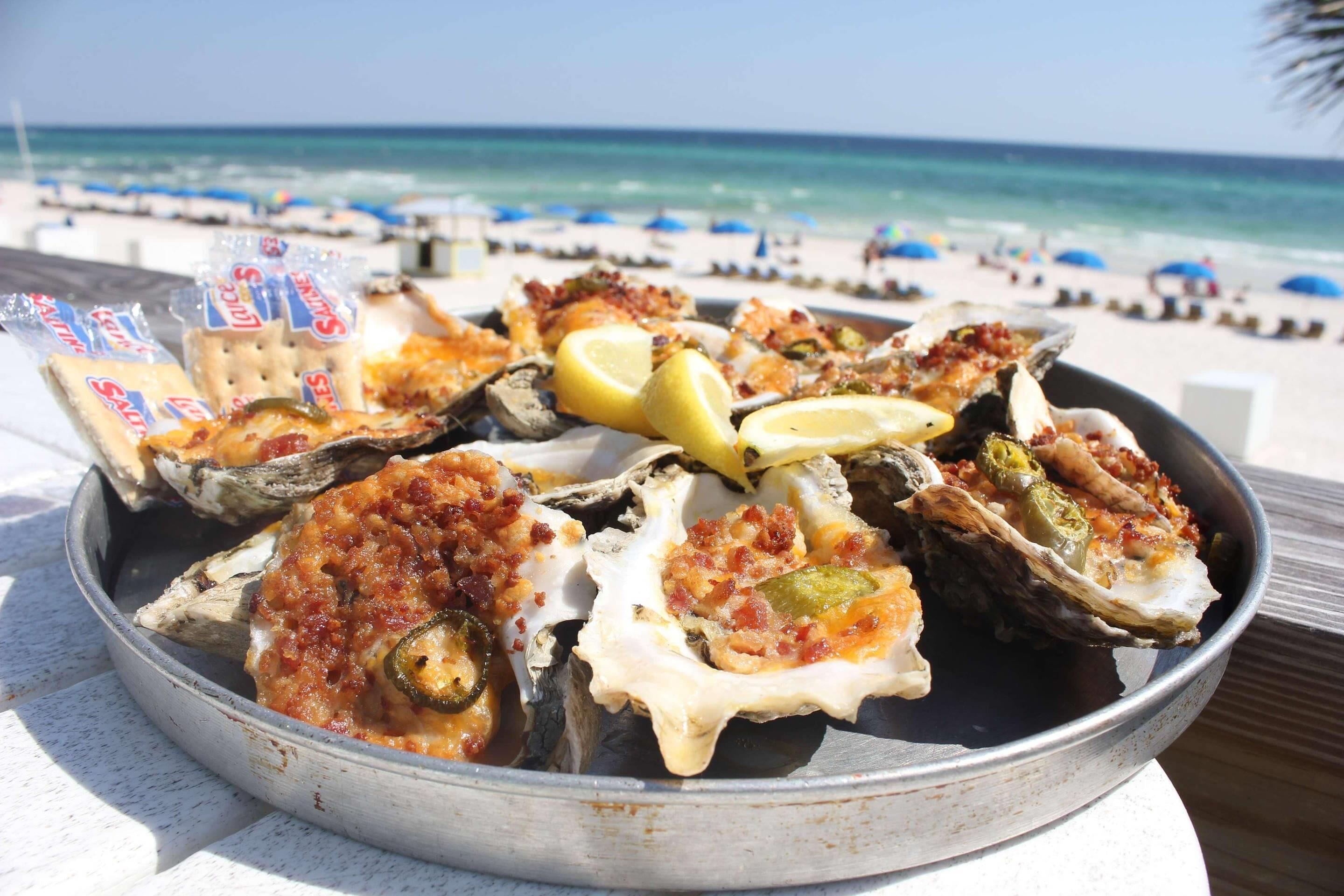 Un plateau d’huîtres servies sur leur coquille, accompagné de quartiers de citron et de crackers, le tout présenté sur une plage.