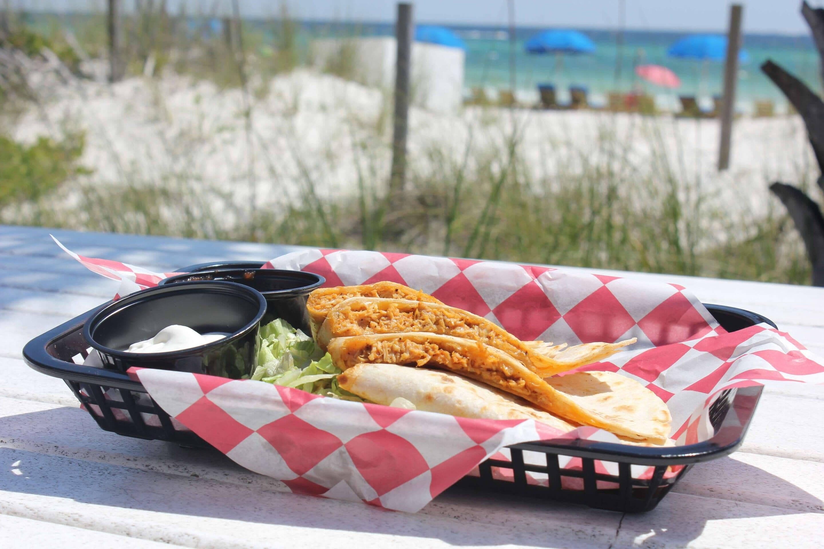 A basket with a sandwich and a small container of sauce on a table with a beach view in the background.