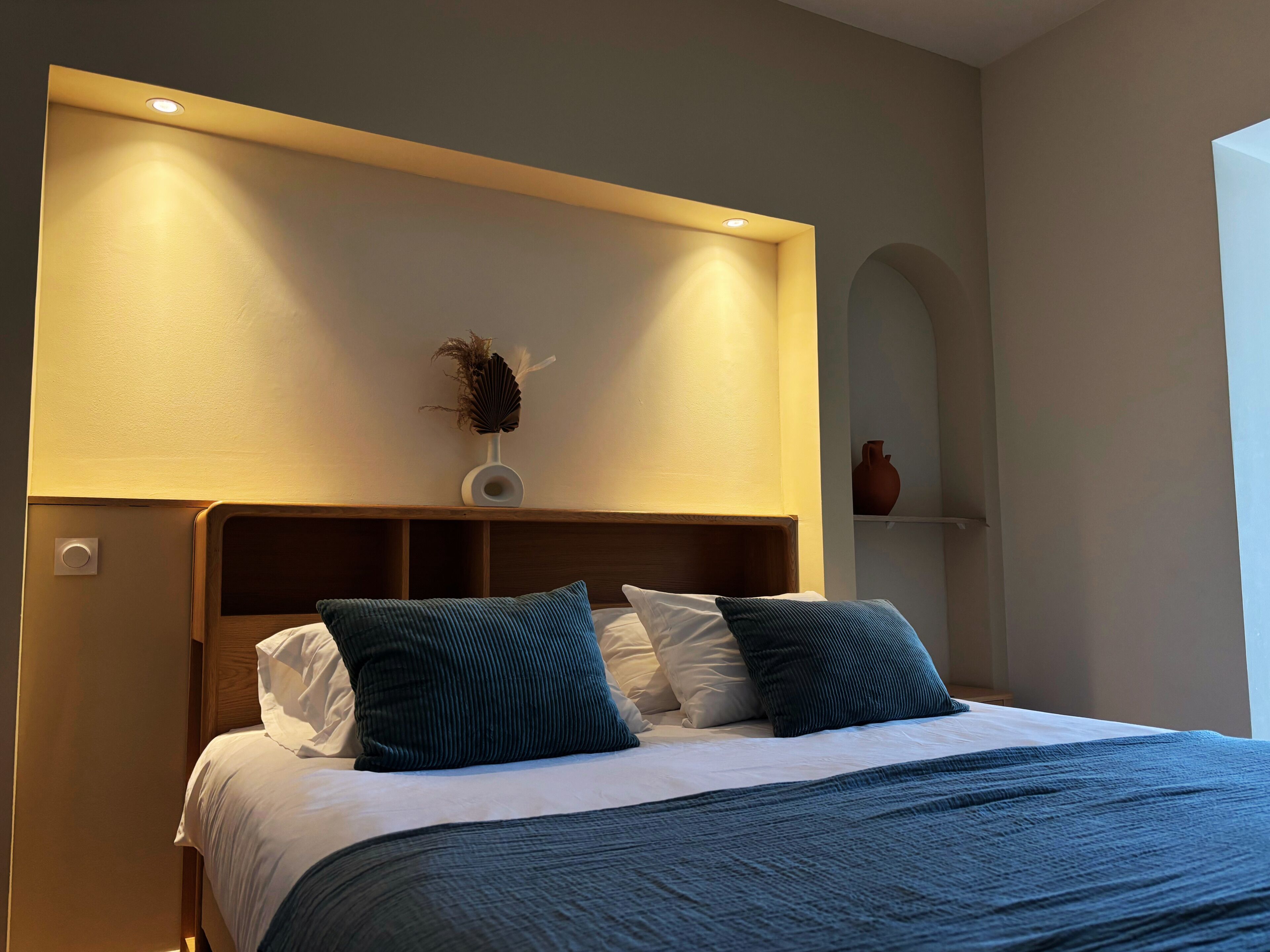 A neatly made bed with a headboard, a vase with dried plants, and a wall-mounted shelf with a vase.