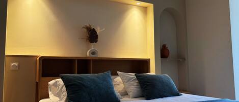 A neatly made bed with a headboard, a vase with dried plants, and a wall-mounted shelf with a vase.