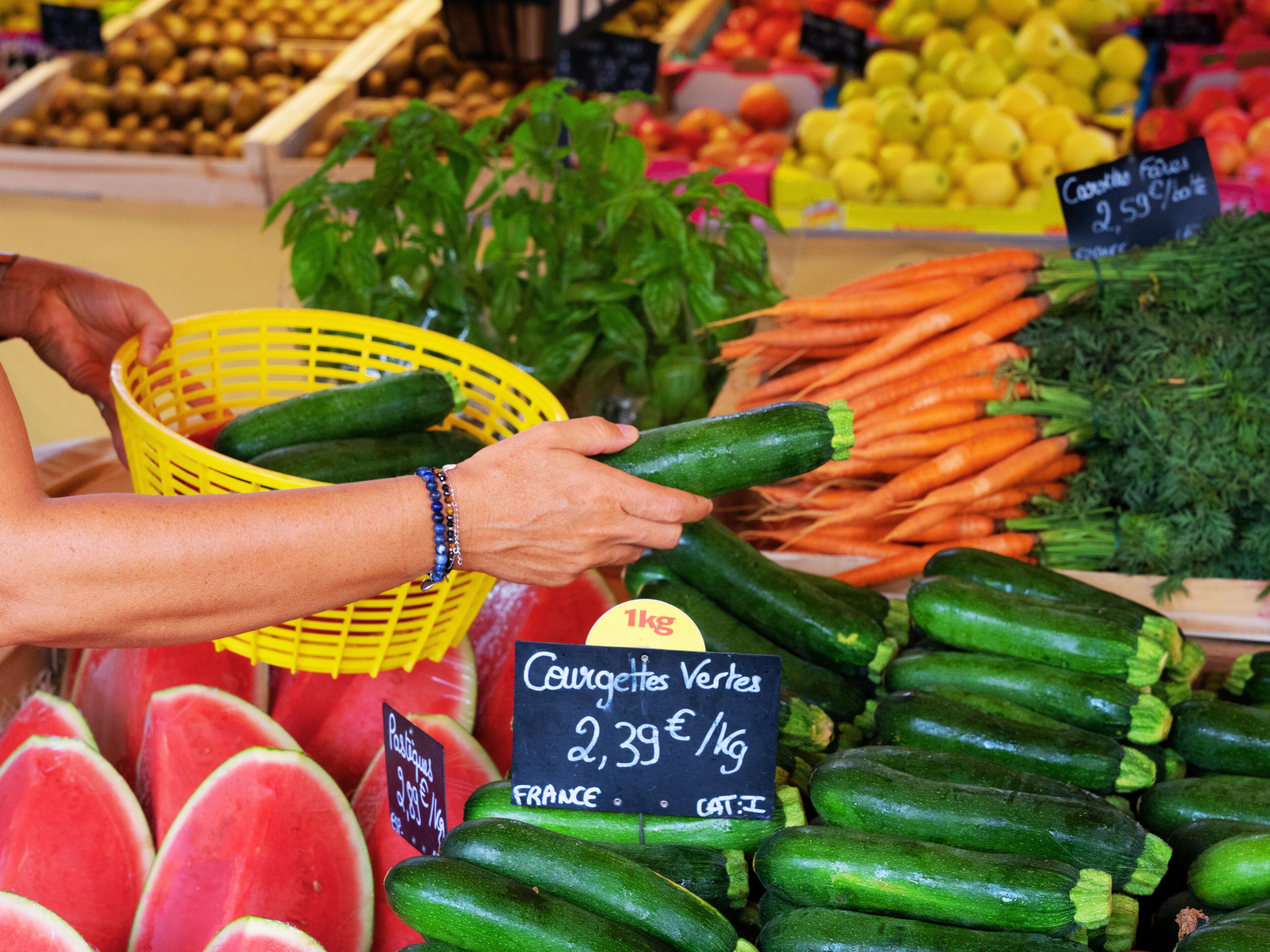 Une personne choisit des concombres sur un étal de marché, avec des étiquettes de prix et d’autres légumes en arrière-plan.