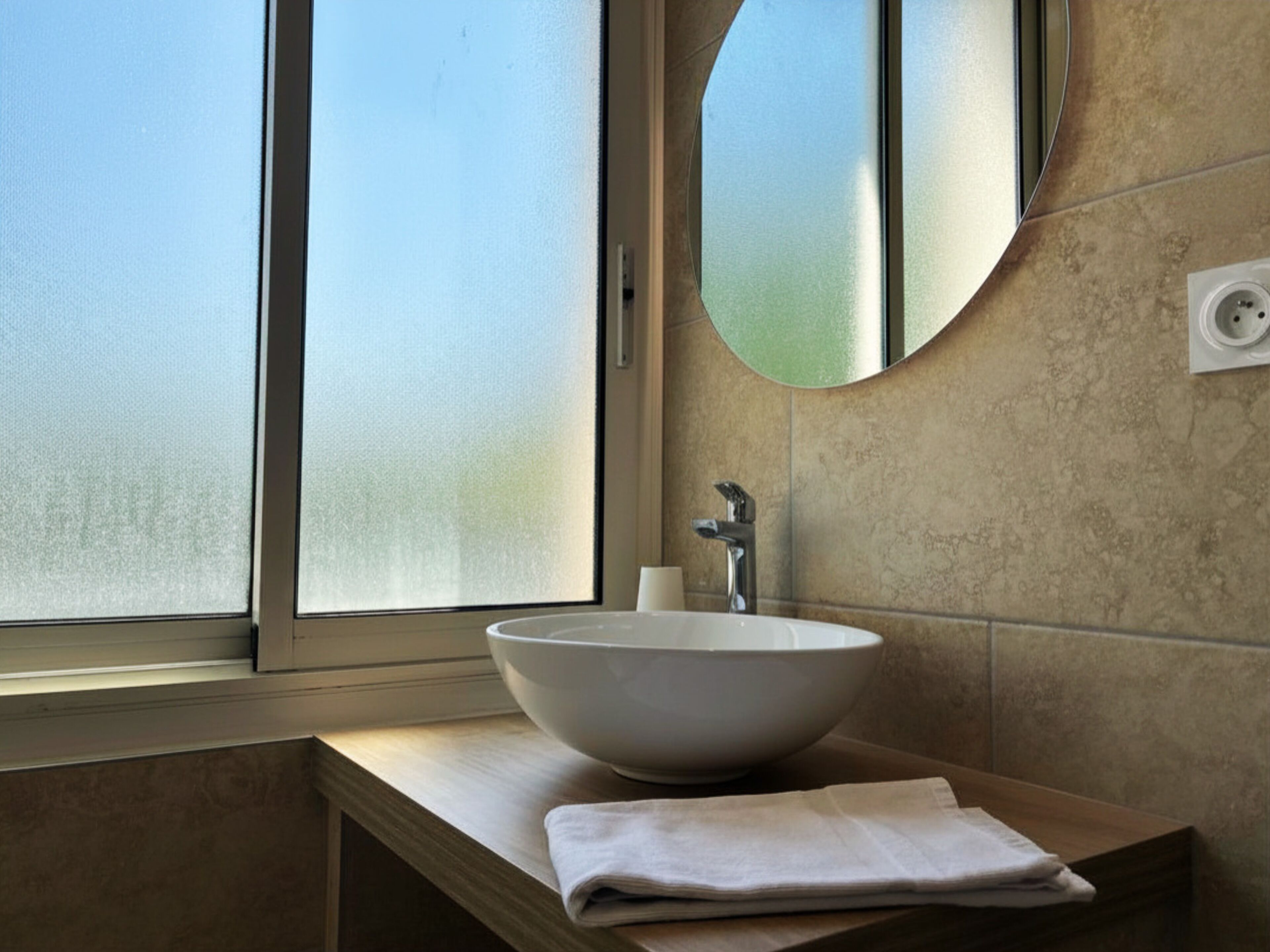 A bathroom with a large window, a white sink, a mirror, and a towel on the counter.