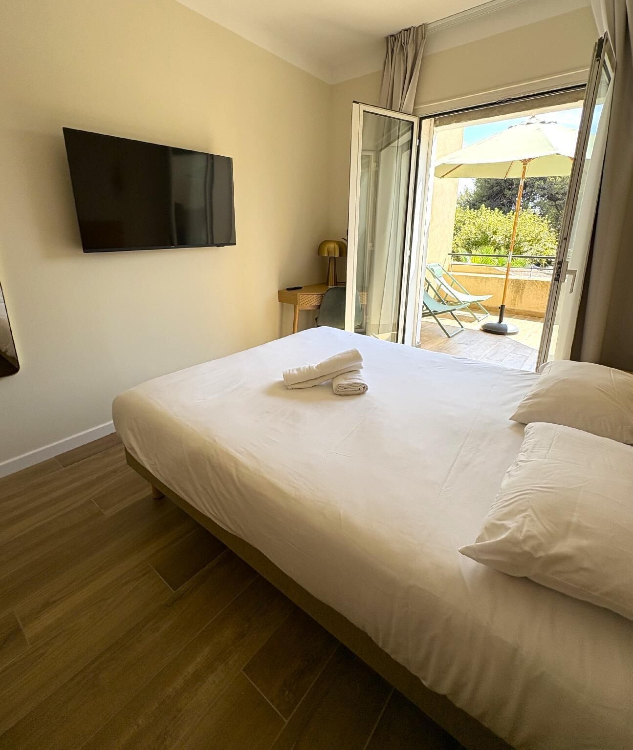Quarto de hotel com cama, televisão, escrivaninha e varanda com mobiliário exterior.