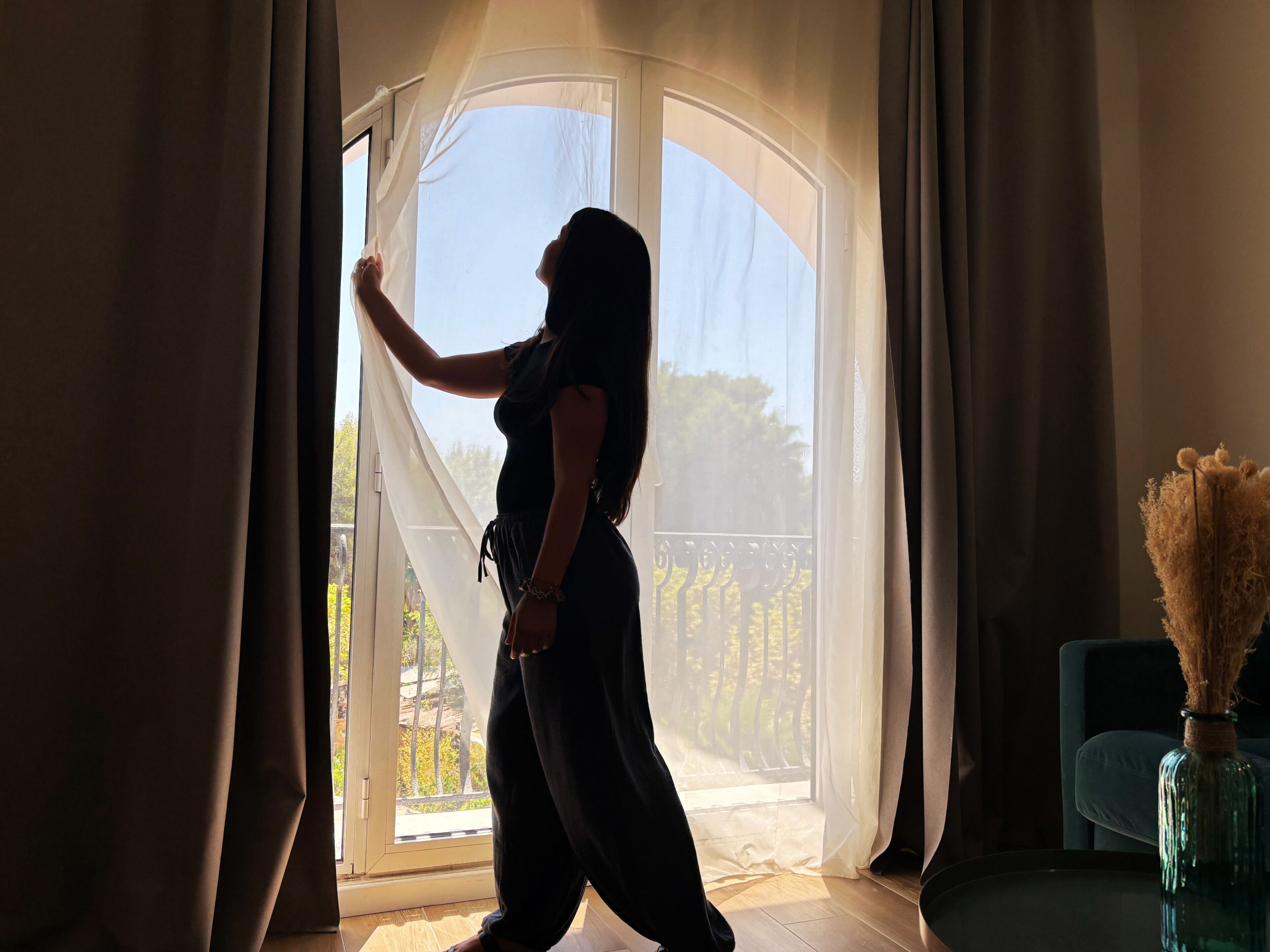 Eine Person steht an einem Fenster mit durchscheinenden Vorhängen, mit einem Balkon und Geländer sowie einer Vase mit Trockenpflanzen auf einem Tisch.
