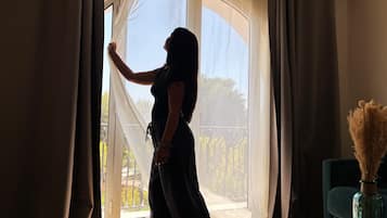 Eine Person steht an einem Fenster mit durchscheinenden Vorhängen, mit einem Balkon und Geländer sowie einer Vase mit Trockenpflanzen auf einem Tisch.