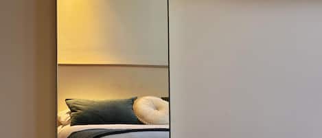 Espejo en la pared que refleja una cama con almohadas y una mesita de noche.