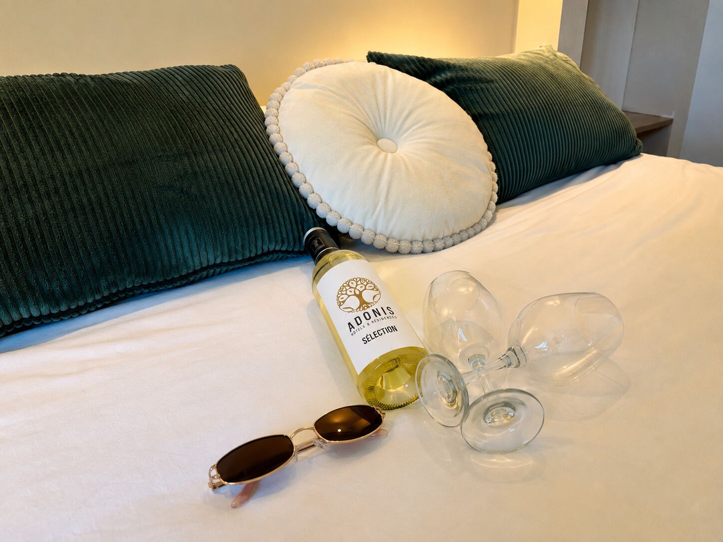 Ein Bett mit grünen Kissen, eine Weinflasche, Weingläser und eine Sonnenbrille.