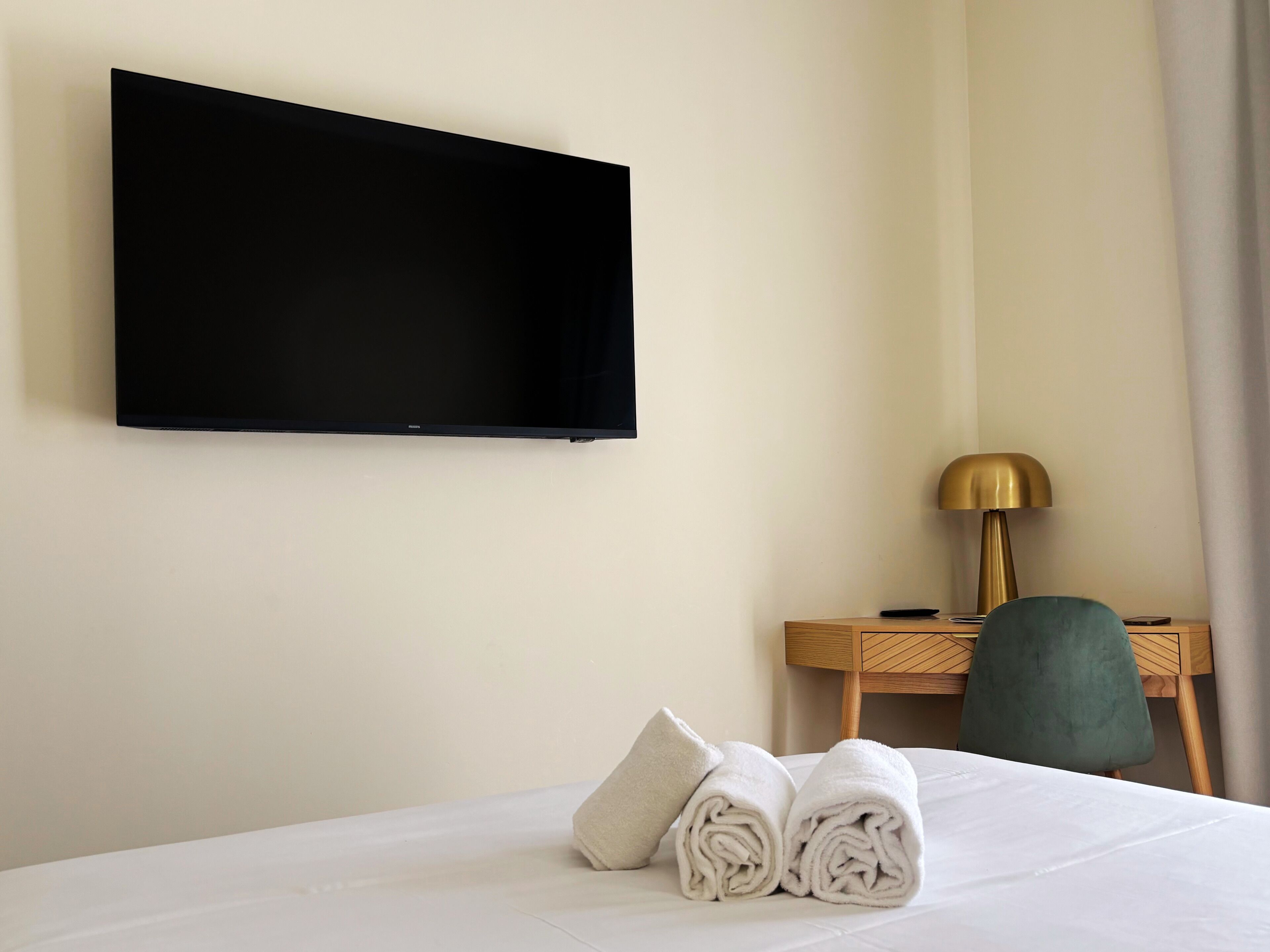 A hotel room with a bed, a flat-screen TV mounted on the wall, a desk with a lamp, and a chair.