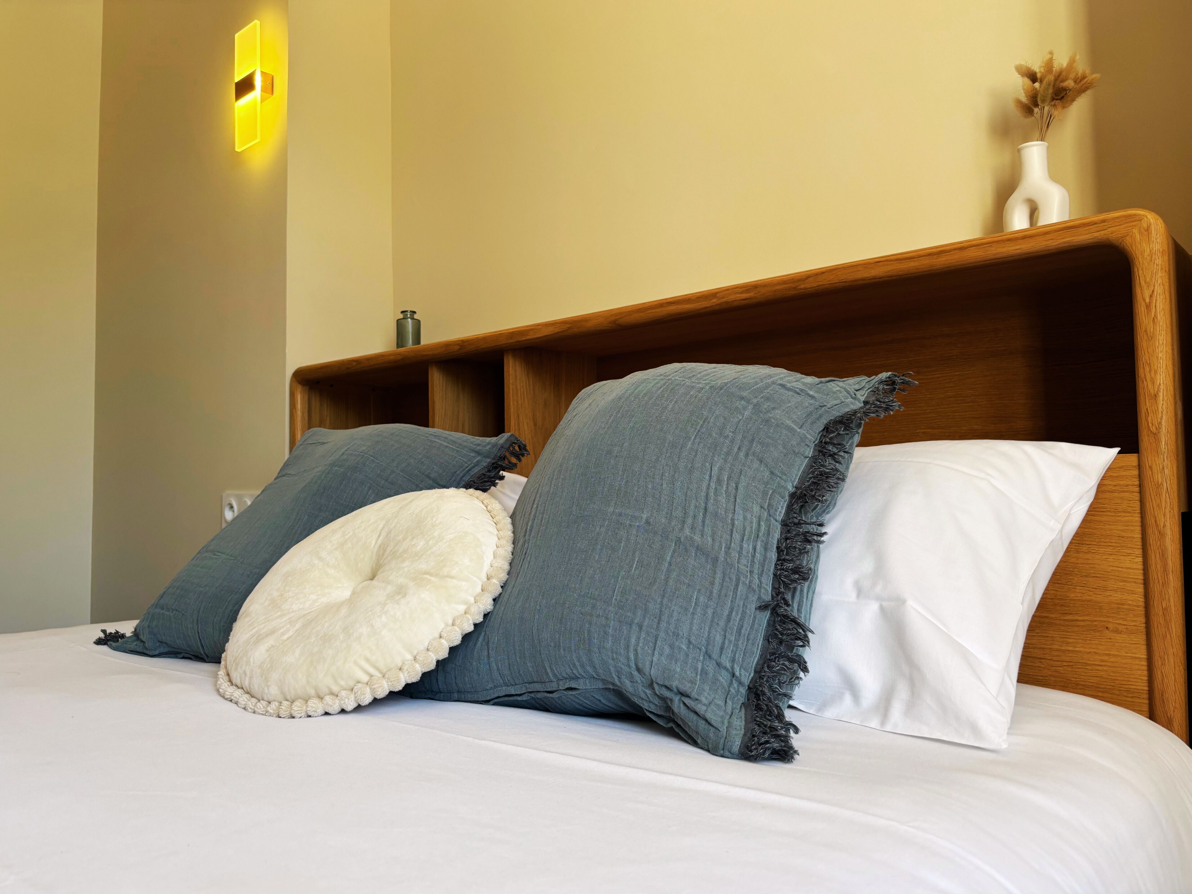 A bed with a wooden headboard, two pillows, and a vase with dried flowers on the nightstand.