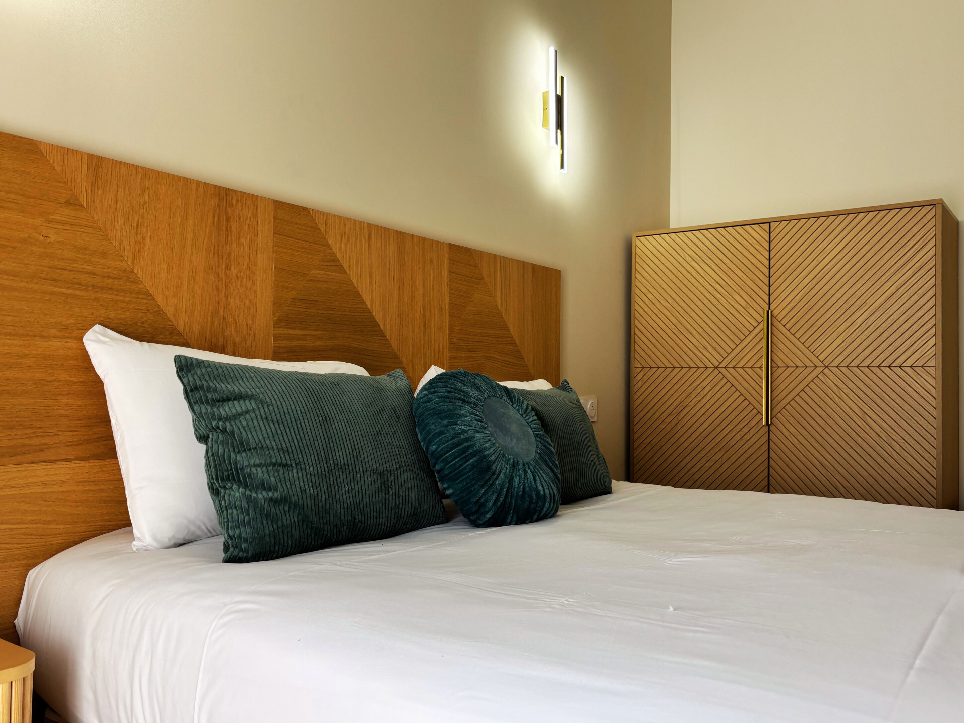 Une chambre d’hôtel avec un lit, une tête de lit en bois, une lampe fixée au mur et une armoire ornée d’un motif géométrique.