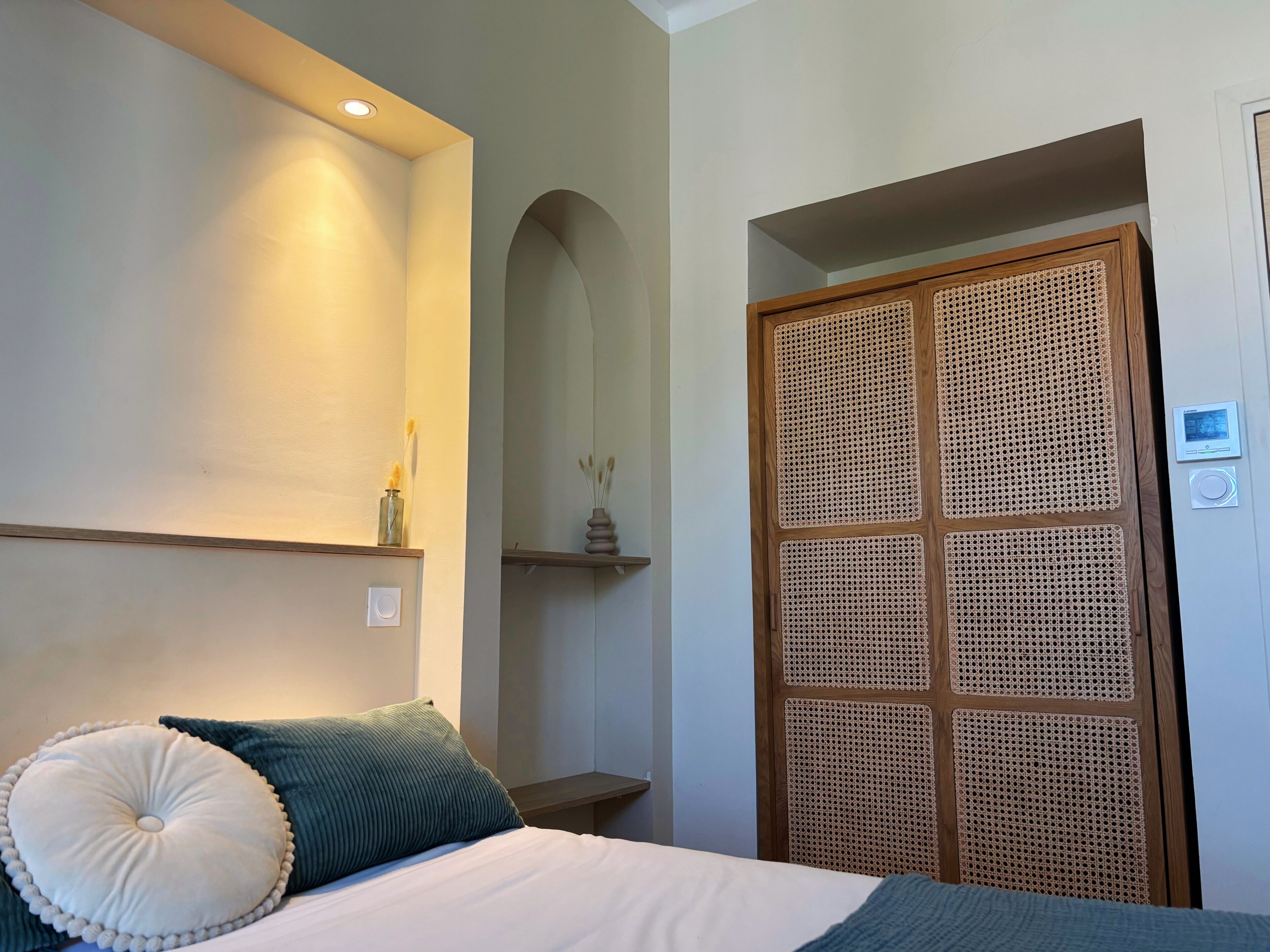 Un dormitorio con cama, almohadas, mesita de noche, estante en la pared, puerta con panel tejido y termostato.