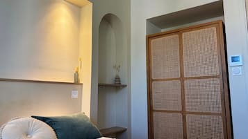 Un dormitorio con cama, almohadas, mesita de noche, estante en la pared, puerta con panel tejido y termostato.