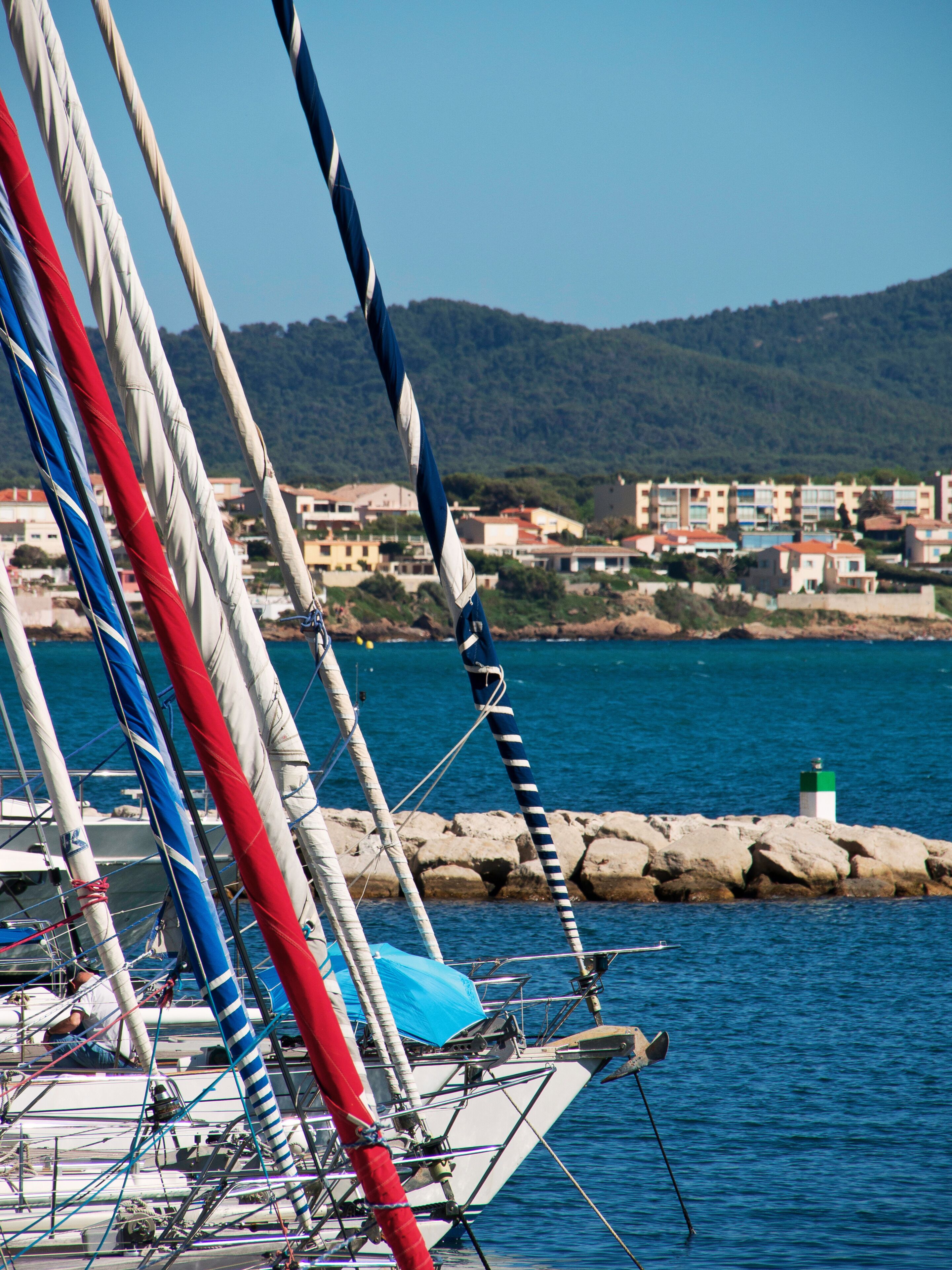 A sailboat with multiple masts and sails, docked near a rocky shoreline with buildings in the background.