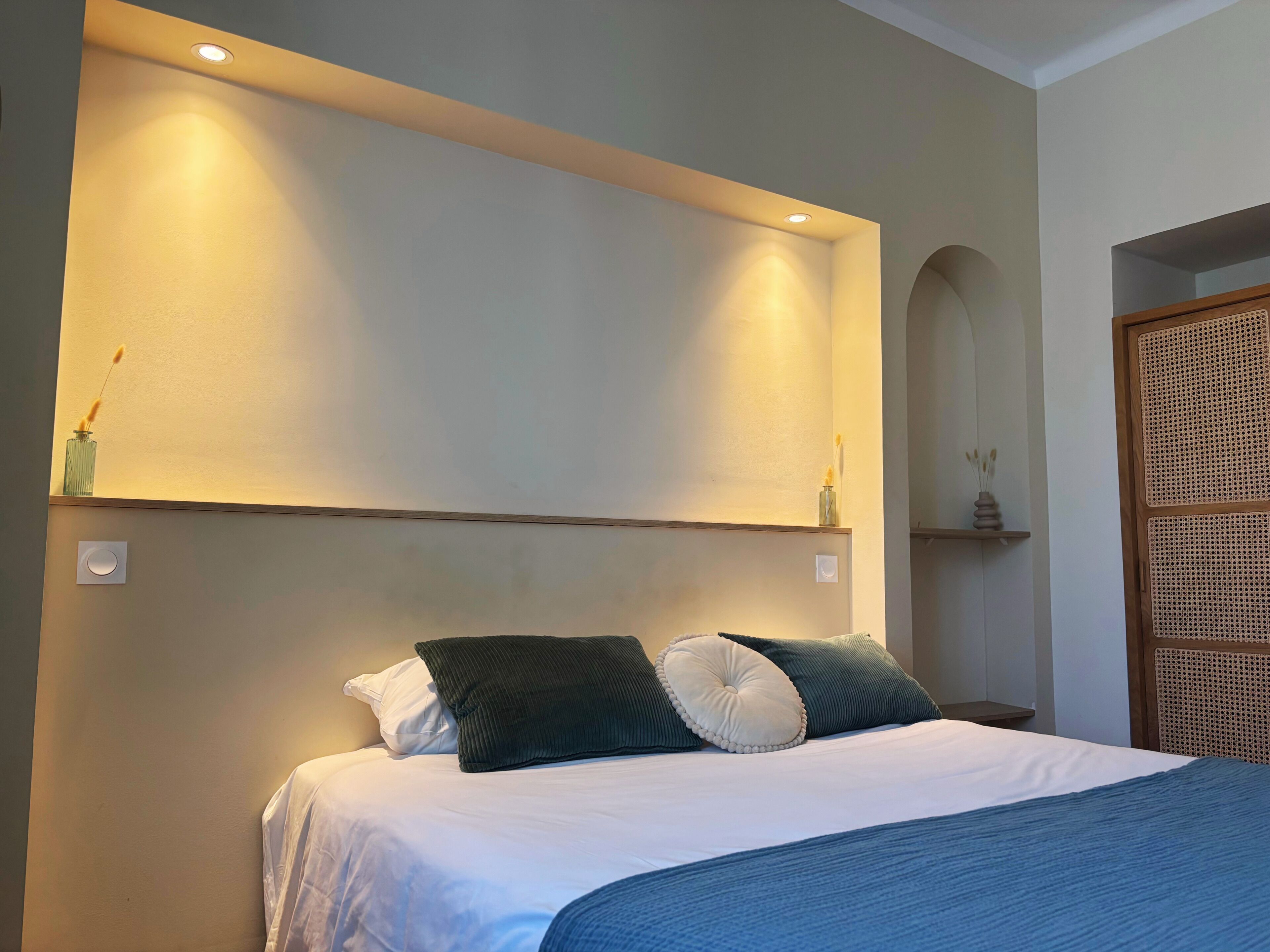 A neatly made bed with white and blue bedding, a wooden headboard, and two wall-mounted sconces providing ambient lighting.