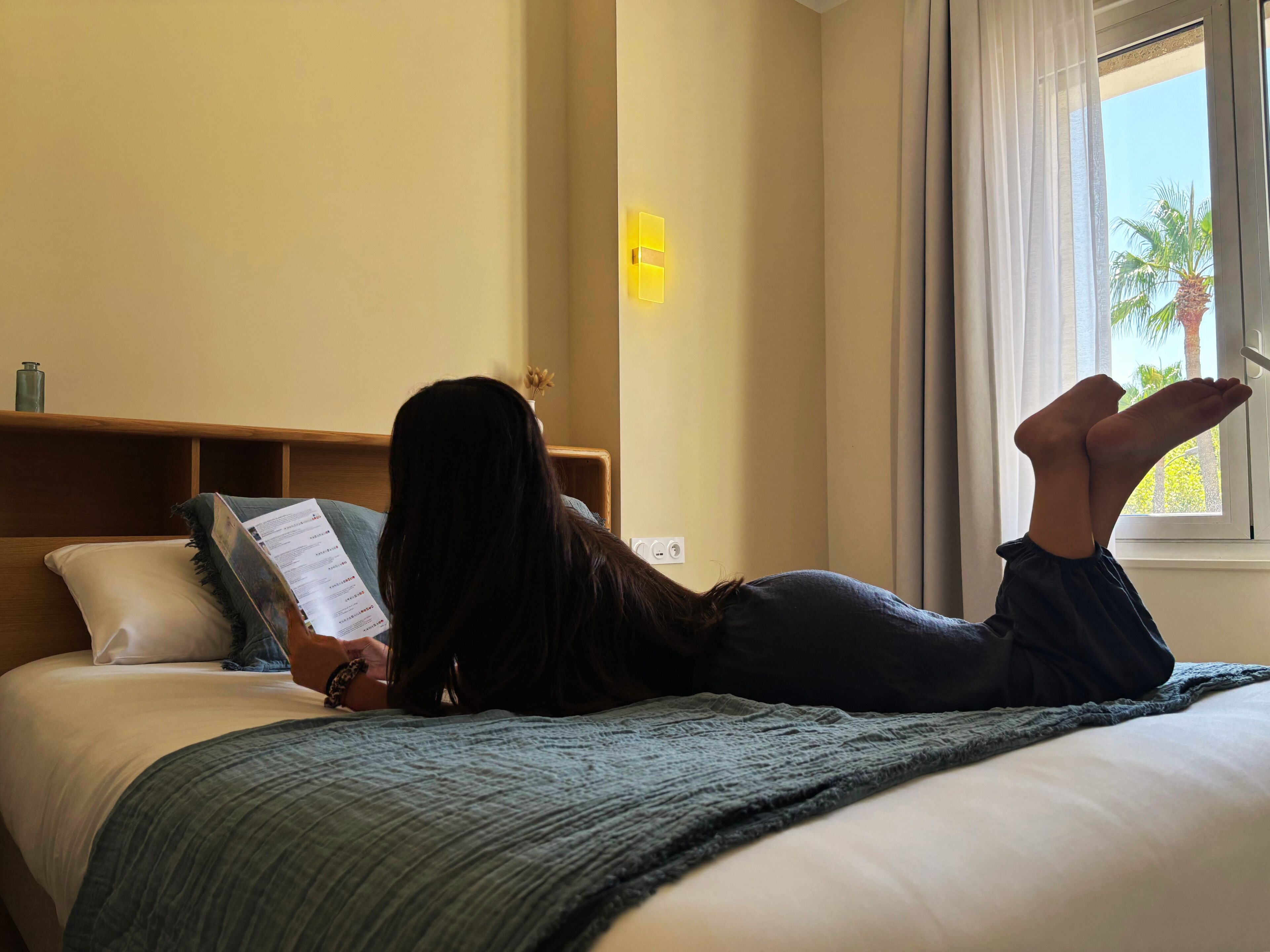 Eine Person liegt auf einem Bett mit einem Kopfkissen, liest ein Buch, in einem Zimmer mit Fenster und Wandlampe.