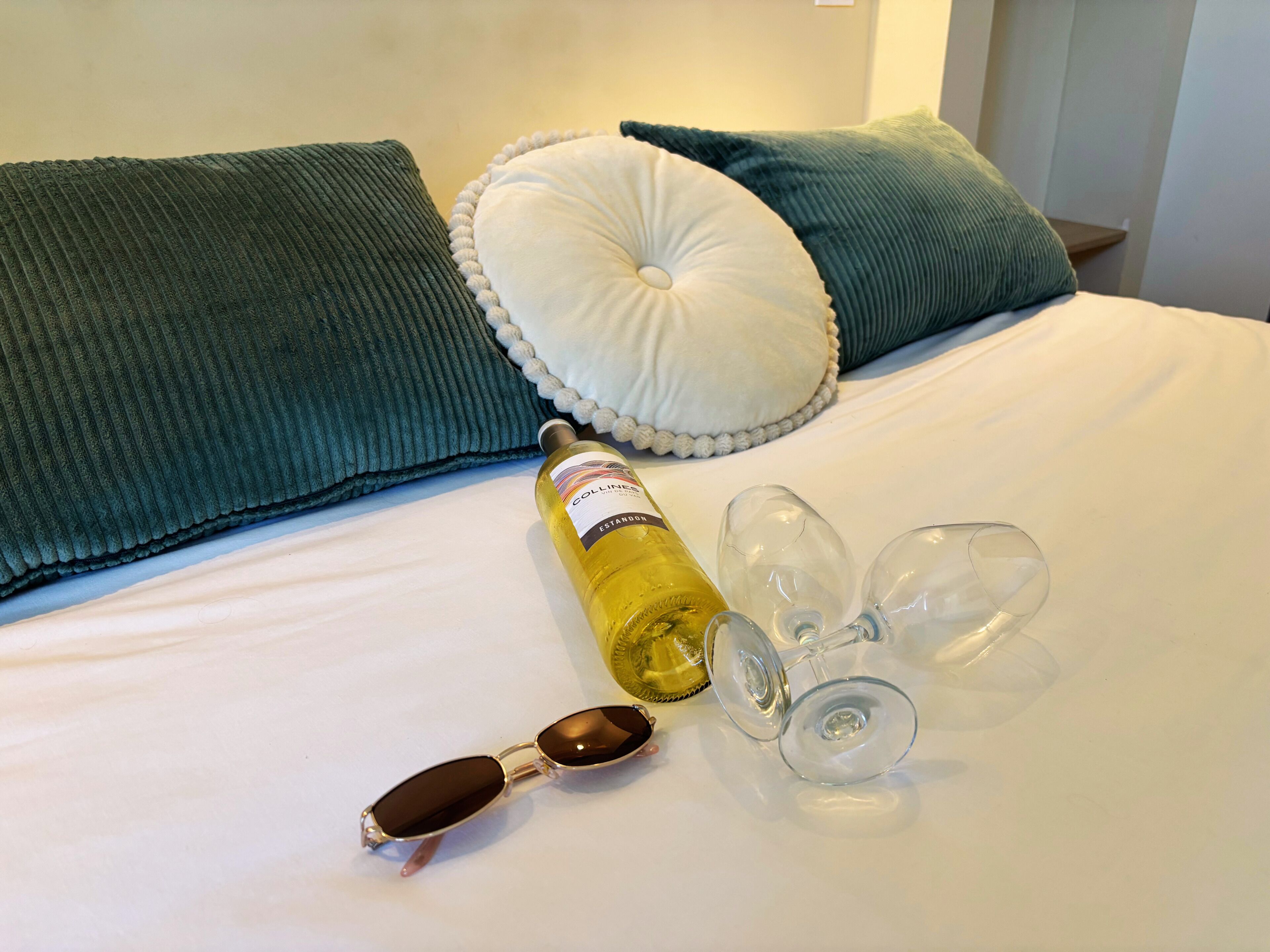 A bed with pillows, a bottle of wine, and wine glasses.