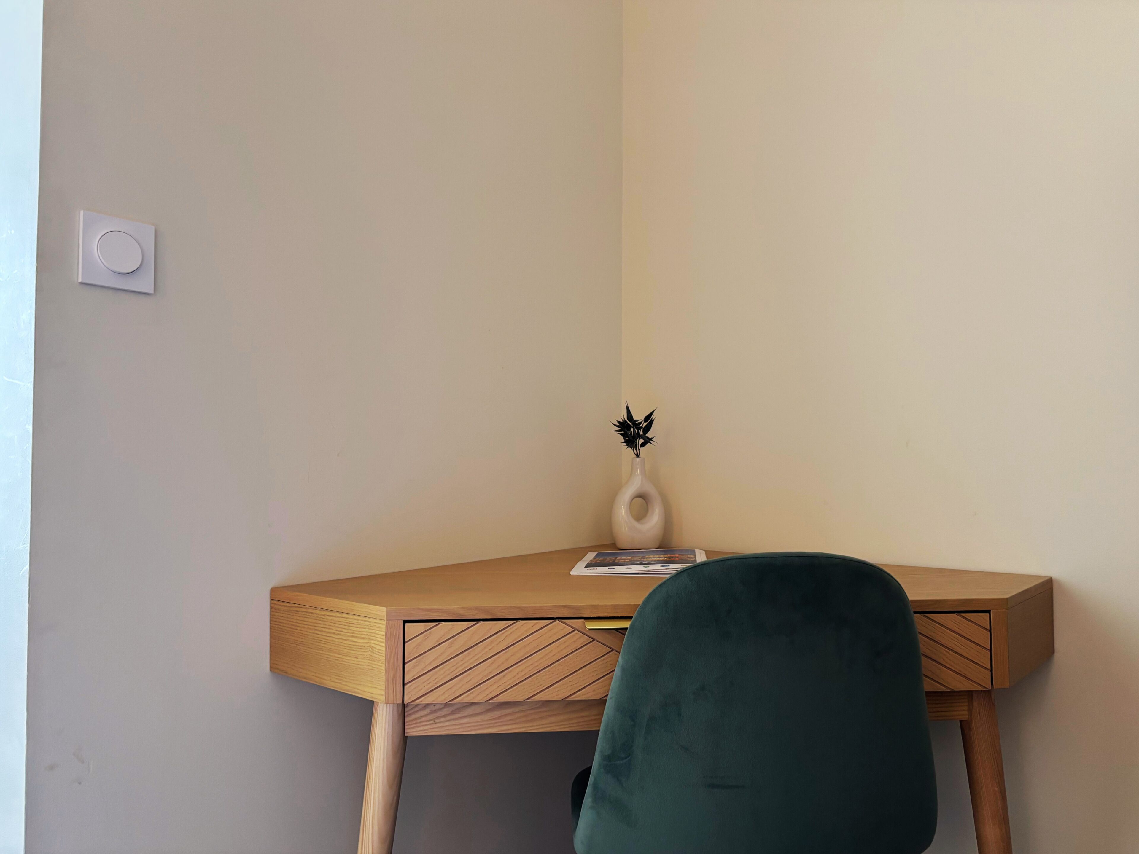 A corner desk with a vase and a book, a green chair, and a light switch on the wall.