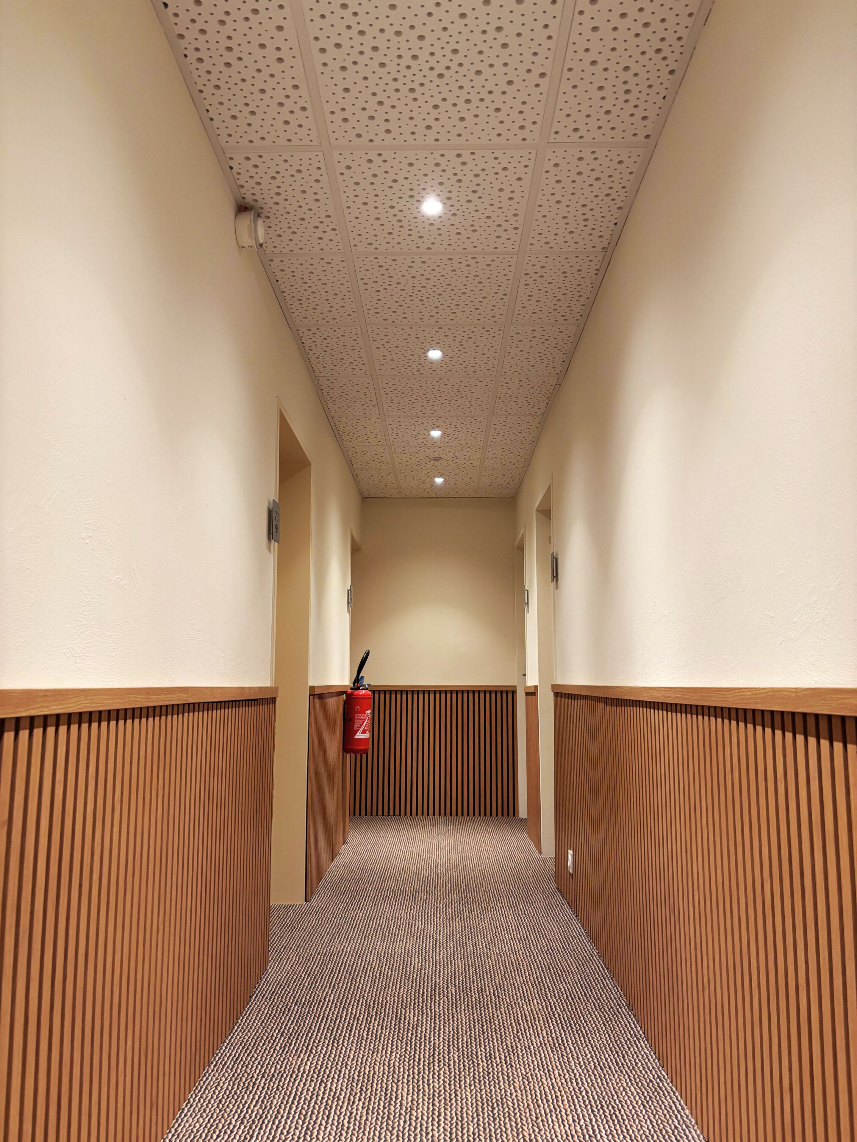 Un couloir doté d’un sol à motifs, de lambris en bois sur les murs, d’un plafond avec des panneaux perforés et d’un extincteur fixé au mur.