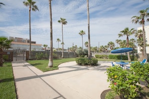 Zona residencial con un camino pavimentado, palmeras, un patio cercado y mobiliario exterior con una sombrilla azul.