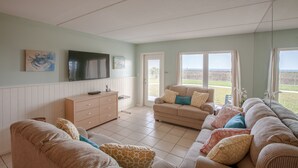Een ruime woonkamer met een grote zithoek, een televisie en uitzicht op het buitengebied door de ramen.