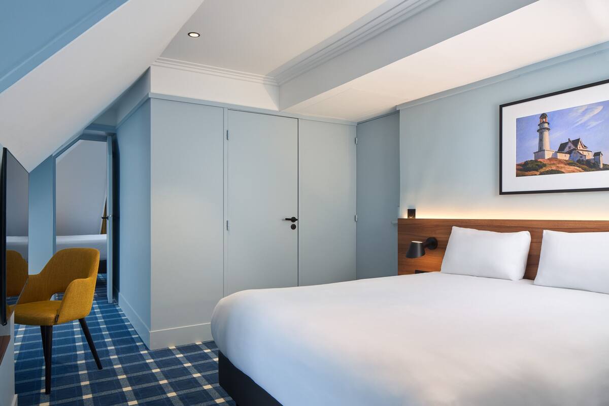 A hotel room with a large bed, a chair, a wardrobe, and a painting on the wall.