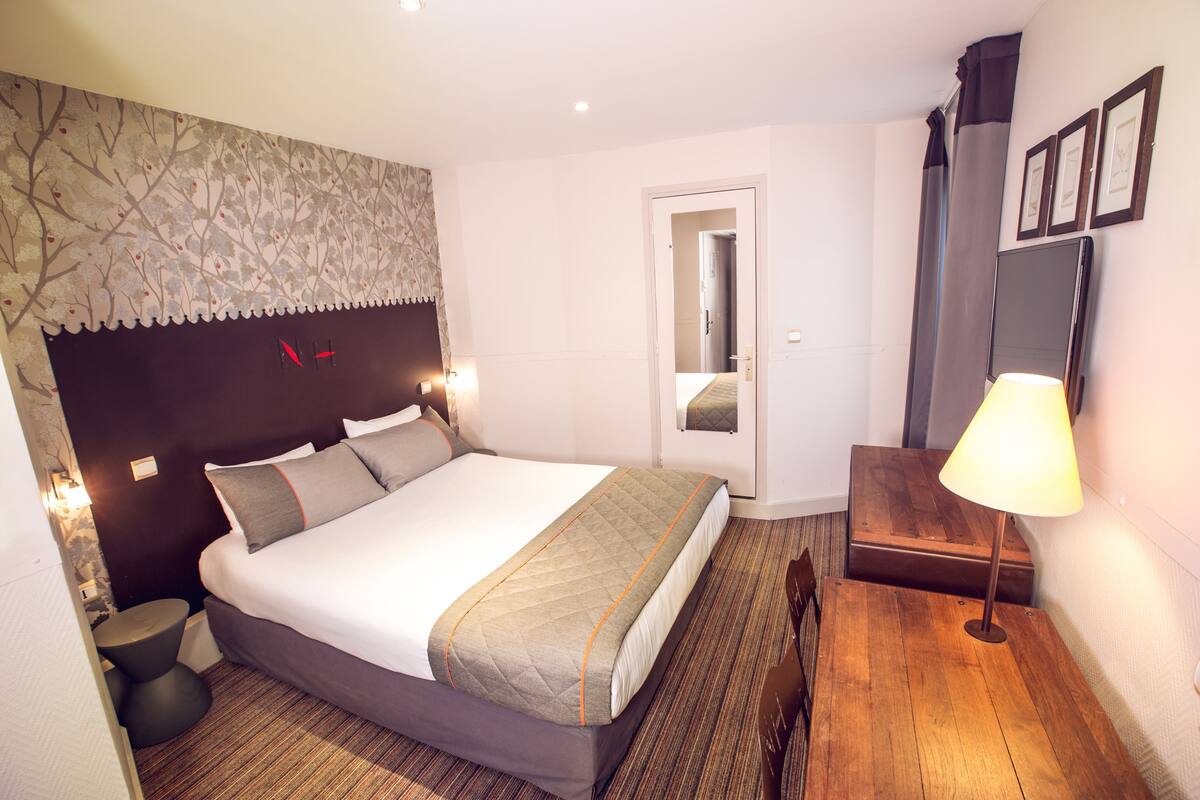 A hotel room with a bed, bedside tables, a lamp, a mirror, and a wall with framed pictures.