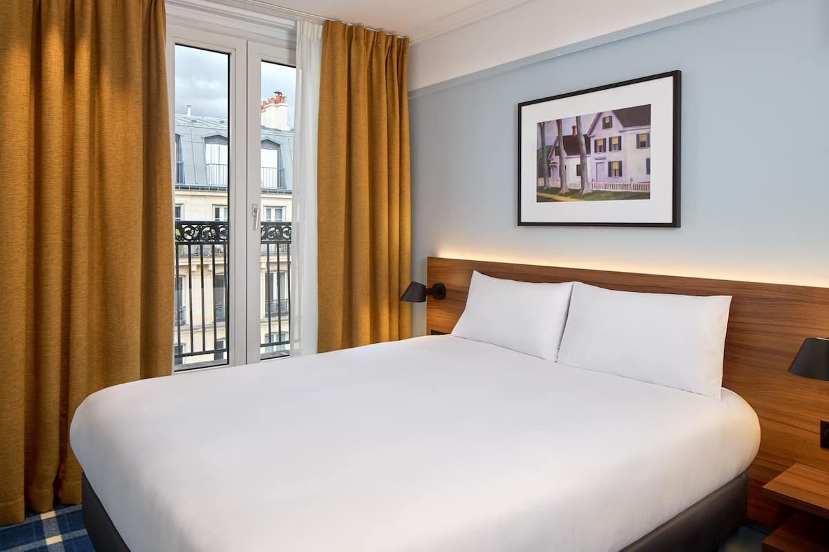 A hotel room with a large bed, a wooden headboard, a framed picture on the wall, and a view of the city through the window.