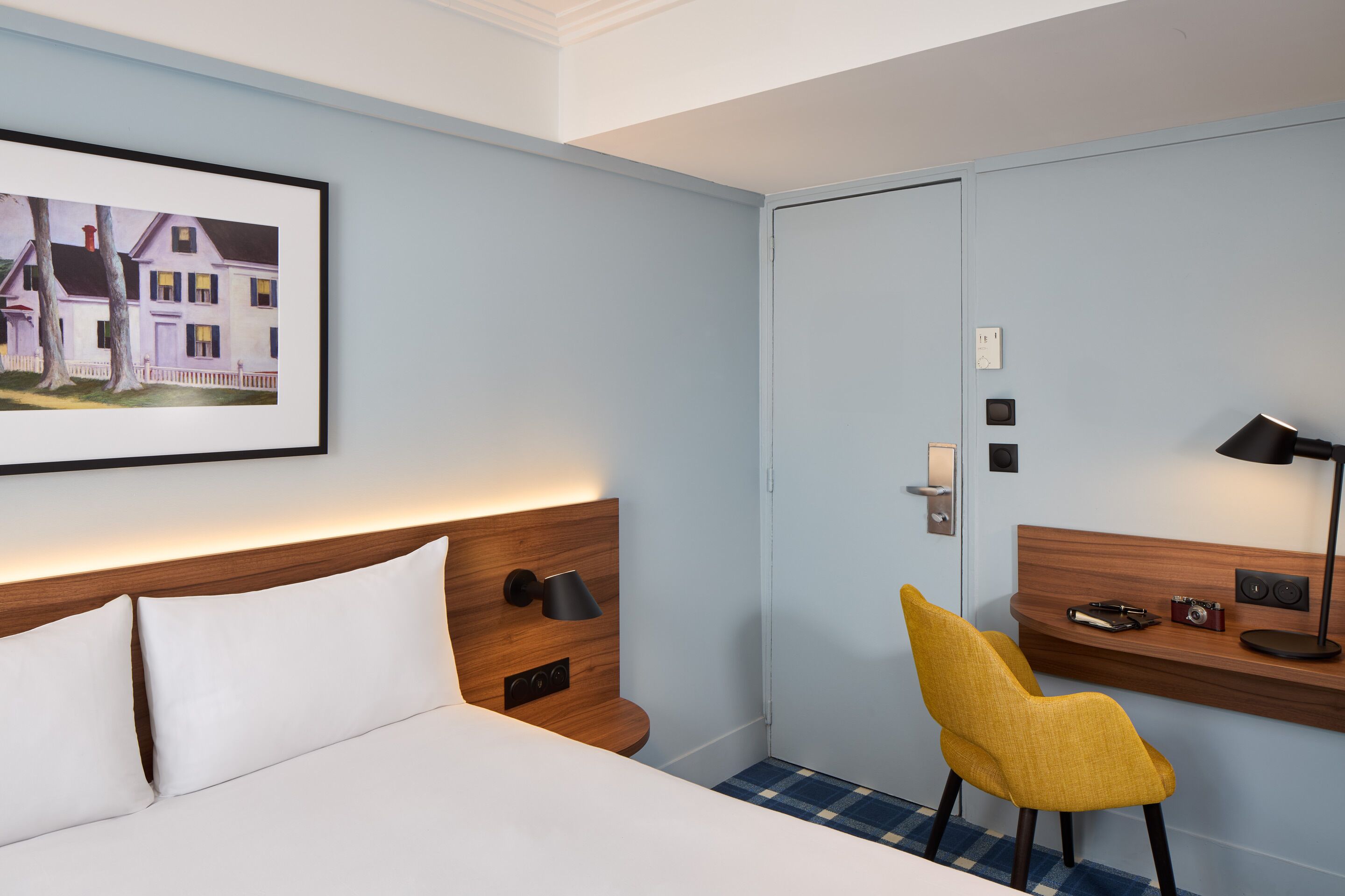 Habitación de hotel con cama, escritorio y silla, un cuadro en la pared y una puerta.