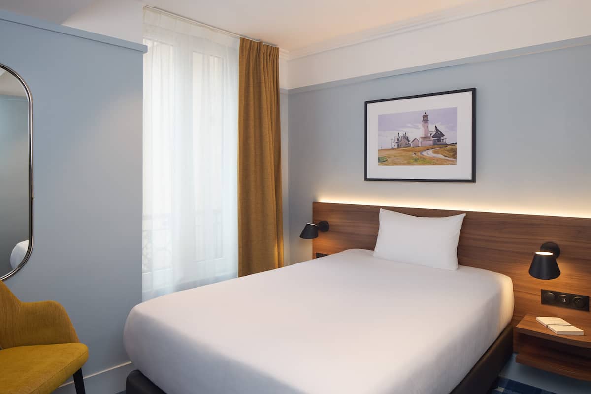A hotel room with a large bed, a wooden headboard, a nightstand with a lamp, a framed picture on the wall, and a window with curtains.